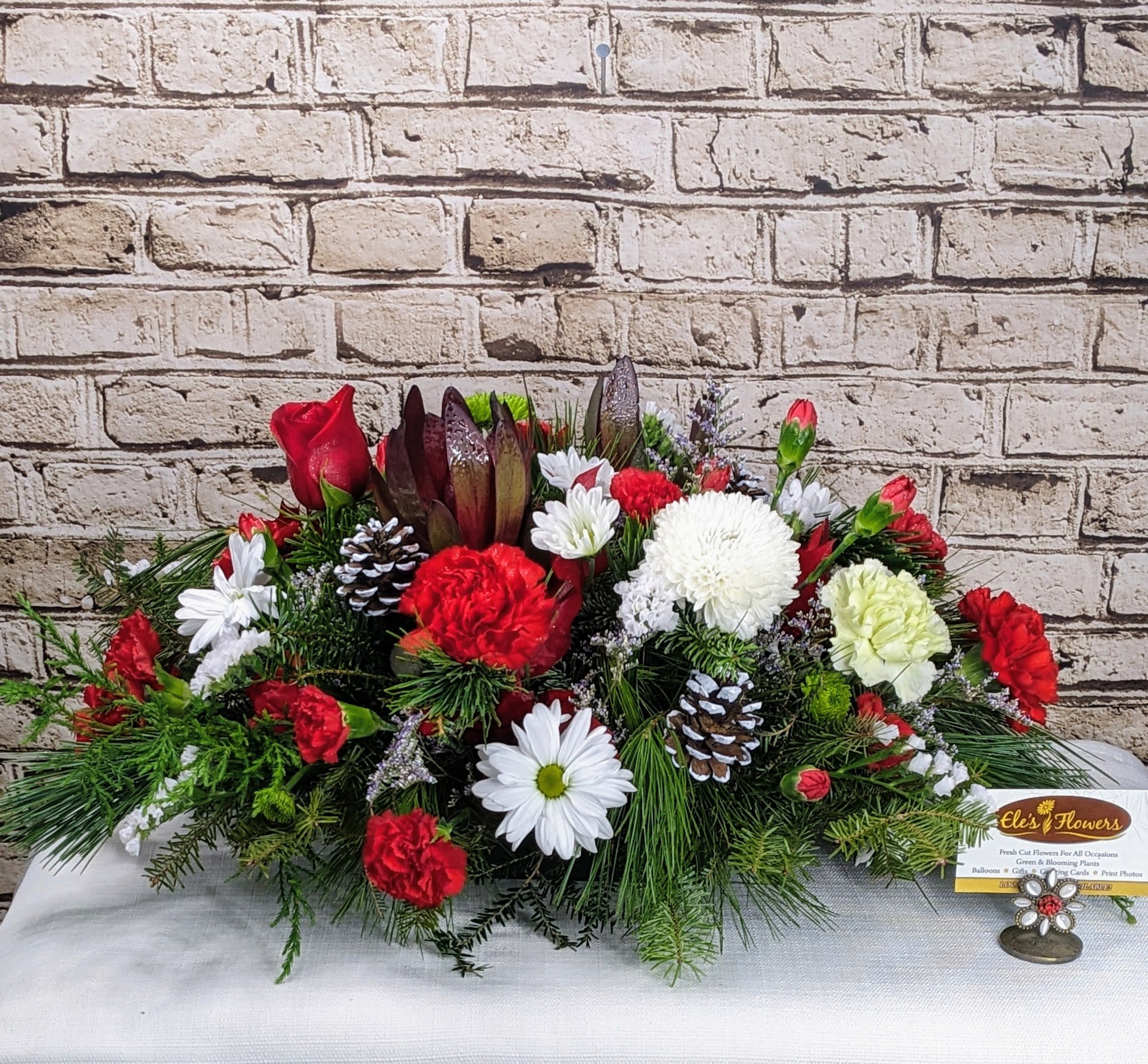 Fireside Cheer Centerpiece - Bursting with festive color and cozy holiday charm, Fireside Cheer is a joyful centerpiece designed to bring warmth and happiness to your holiday table. Rich reds and crisp whites pop beautifully against layers of fresh, textured evergreens, while natural woodland accents add a touch of rustic magic. Full, lush, and undeniably merry, this arrangement feels like gathering around a crackling fire with loved ones—perfect for celebrating the season, hosting holiday meals, or sharing a little Christmas joy with someone special.  Approximate Dimensions: 22"L x 14"H