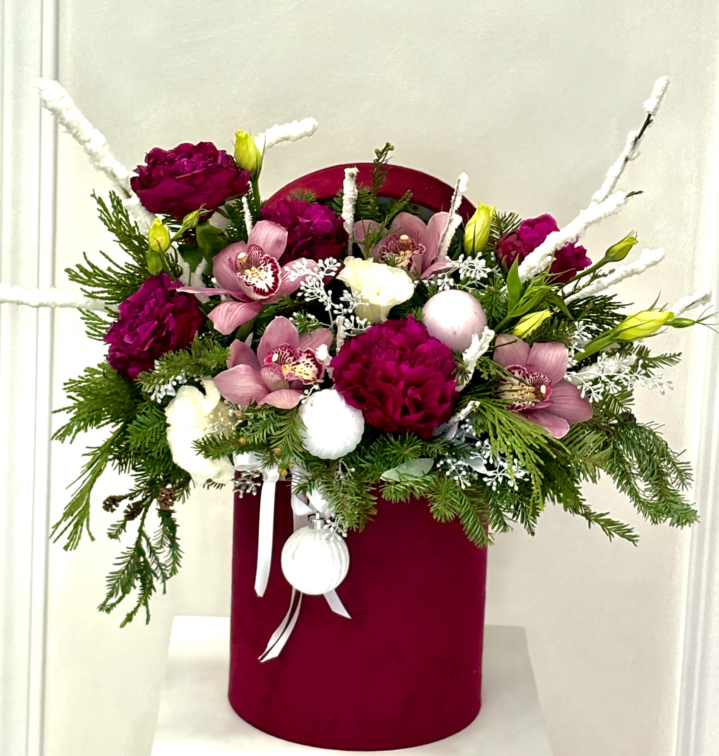 Winter Berry Elegance Hat Box By VLM Flowers - A rich and festive arrangement featuring deep burgundy blooms, soft pink cymbidium orchids, and creamy white accents, all nestled in lush winter evergreens. Designed in a velvet burgundy hatbox and finished with delicate frosted branches and holiday ornaments, this arrangement brings refined warmth and seasonal charm—perfect for Christmas gifting or elegant winter décor.