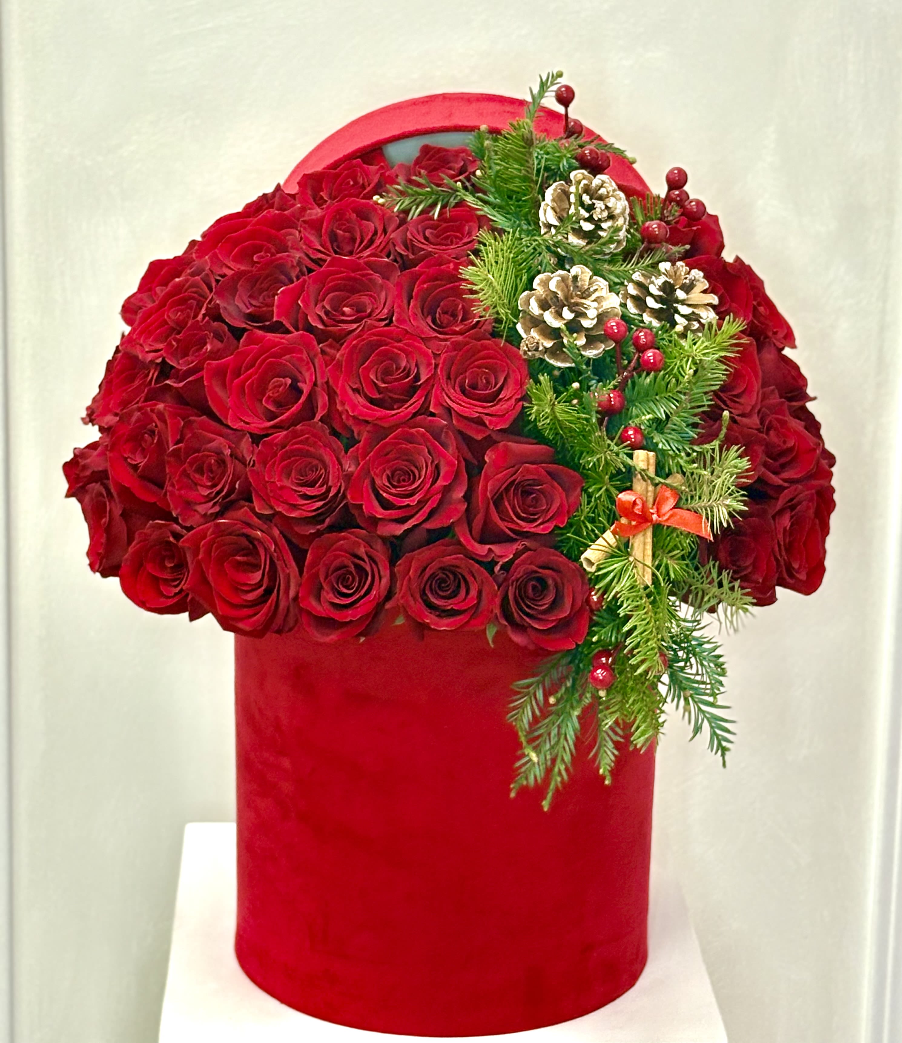 Crimson Holiday Luxe Hat Box By VLM Flowers - A striking dome of velvety red roses arranged in a bold red hatbox, accented with fresh evergreen branches, pinecones, and festive red berries. This luxurious holiday design blends classic romance with Christmas warmth, making it a perfect statement piece for seasonal celebrations, elegant gifting, or festive home décor.