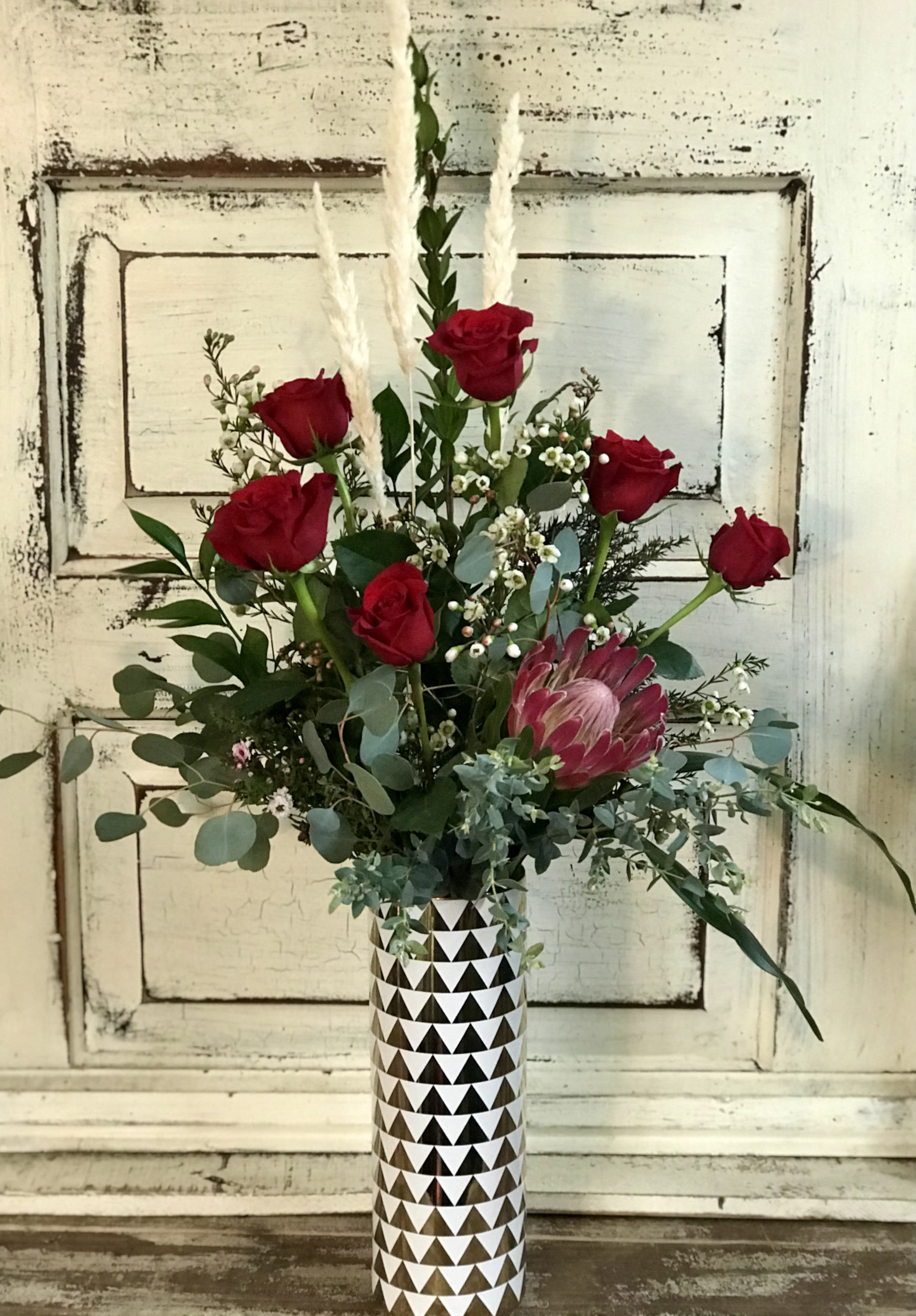 Roses &amp; Protea Vase - A unique ceramic vase contains a half dozen red roses with a tropical protea focal point. A variety of foliage, fillers and pampas grasses complete the dramatic and romantic look.