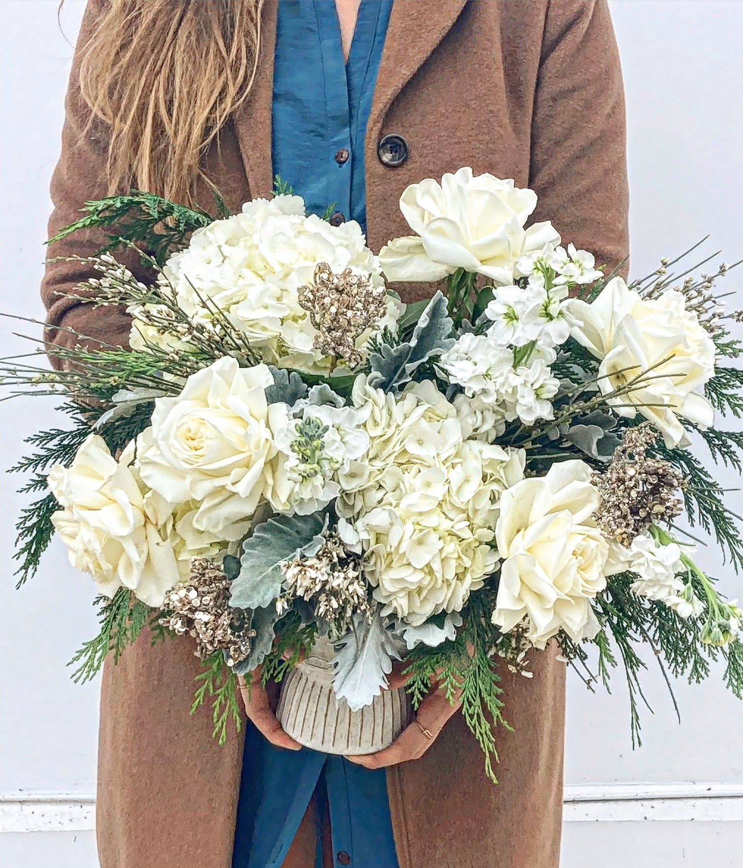 The Sleigh Ride - The Sleigh Ride is a luxurious winter wonderland captured in floral form. Featuring voluminous white hydrangeas, delicate white stock, and lush white roses, this arrangement exudes a serene elegance. Fragrant cedar adds a festive, woodsy aroma, while silvery dusty miller lends a frosted, ethereal touch. Accents of airy white genista weave through the design, evoking the gentle beauty of freshly fallen snow. Arranged in a sophisticated snowflake vessel, the Sleigh Ride is the perfect centerpiece to bring a touch of crisp winter magic to your holiday celebrations.