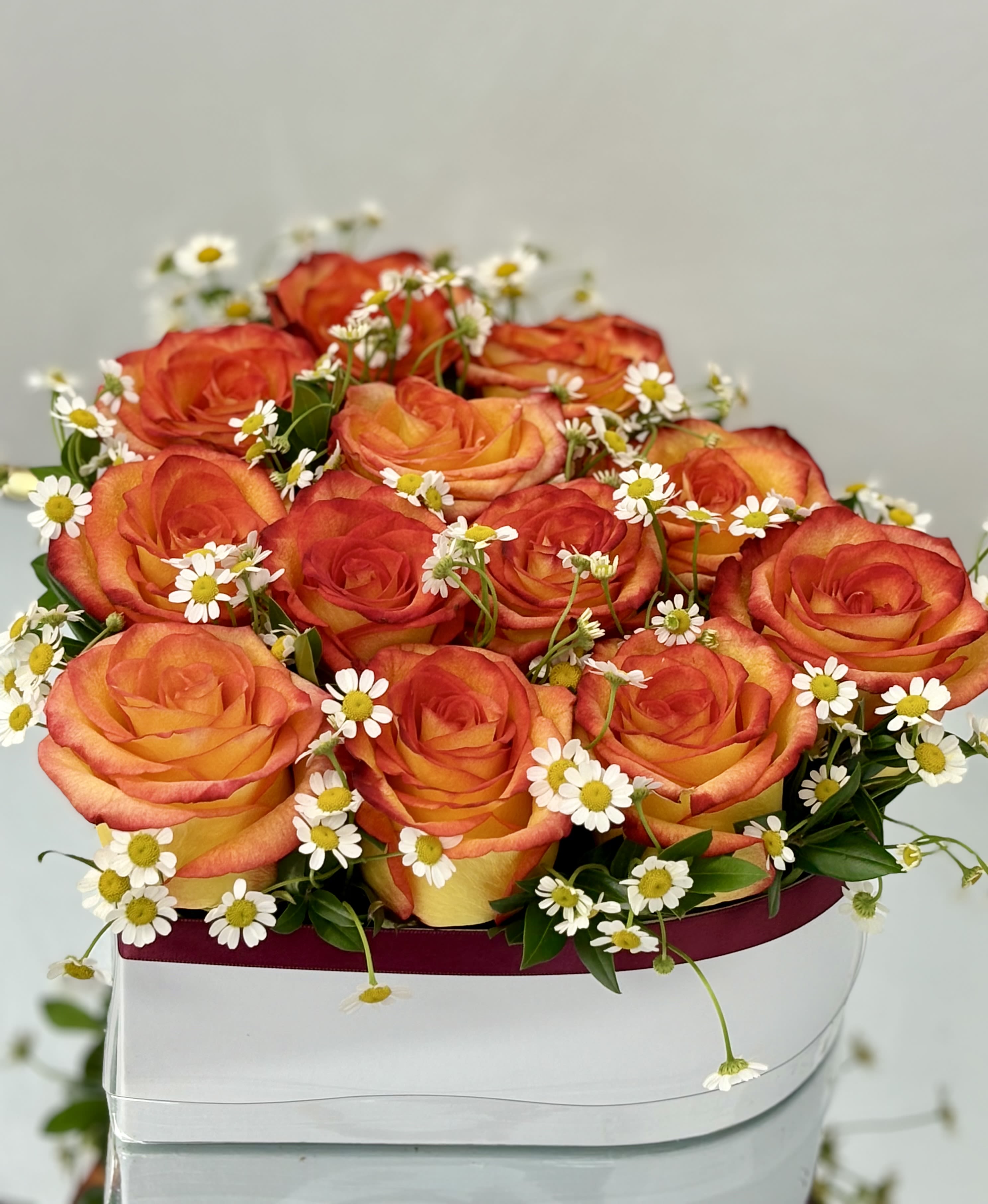 Sunshine Heart By VLM Flowers - This cheerful heart-shaped box bursts with fiery orange-yellow roses and delicate chamomile blooms, symbolizing warmth, joy, and a touch of playfulness. A perfect way to express sunny feelings—whether for a birthday, thank you, or a romantic surprise full of light and affection.