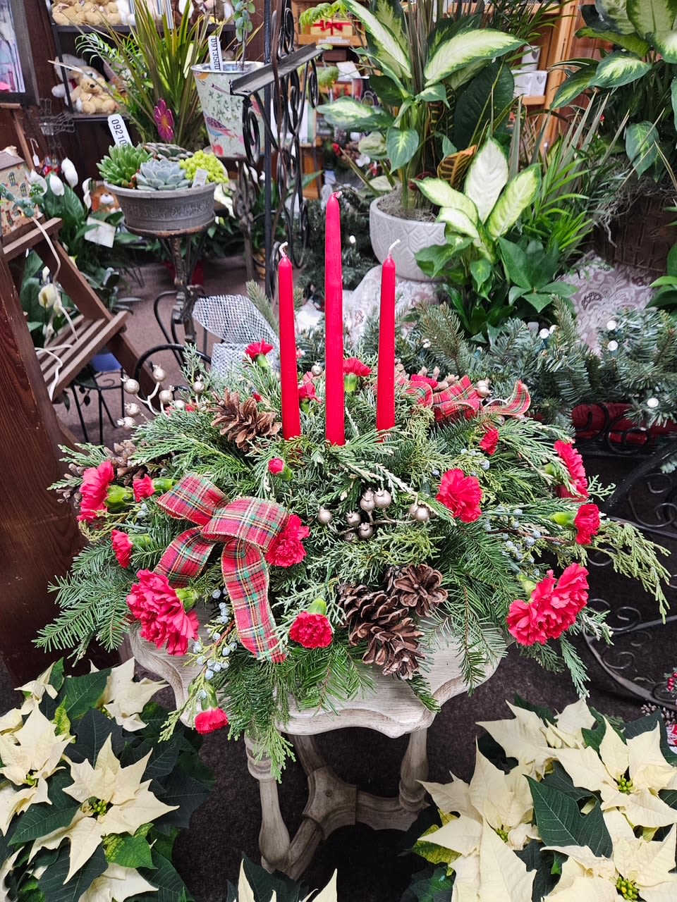 All is Bright - All will be bright this season when you order this joyful Christmas arrangement. A lovely centerpiece will surely light up the holiday festivities beautifully. Miniature carnations are artfully on display with merry touches like shimmery ornaments, pinecones, berries, organza ribbon, and holiday greens. Three graceful red taper candles add the perfect magical touch. Approximately 23" W x 16"