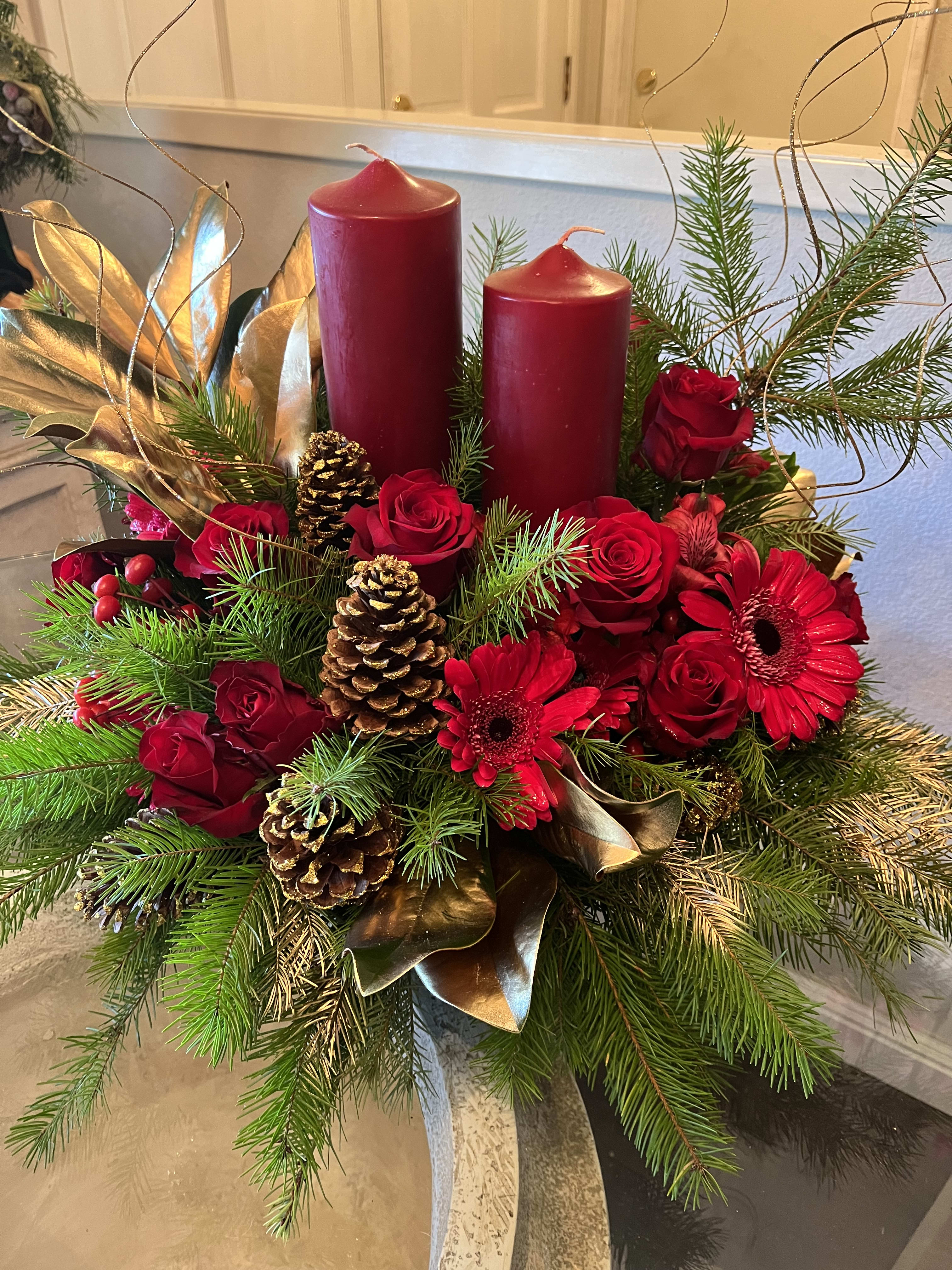 Candlelit Christmas - Ruby red flowers including roses and spray roses are perfectly arranged with golden pinecones and holiday greenery. Two pillar candles, currently available in burgundy, are the perfect touch to light up the night. Approximately 23" W x 18" H
