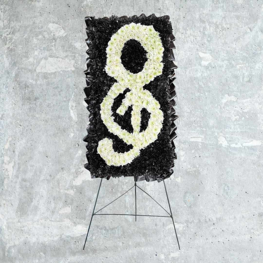 treble Clef - This striking floral treble clef is a custom-designed tribute created to honor a love of music and a life well lived. Crafted on a solid design board, the treble clef is formed entirely from fresh white cushion mums, carefully placed to create smooth curves and beautiful dimension. The bold black background provides strong contrast, allowing the musical symbol to stand out with elegance and clarity.  Thoughtfully designed and handcrafted, this piece requires approximately three hours of detailed floral work from start to finish. The result is a meaningful, artistic memorial that speaks to musicians, music lovers, and anyone whose life was touched by song.  Ideal for funeral and memorial services, this custom floral design is both a visual statement and a heartfelt expression of remembrance.
