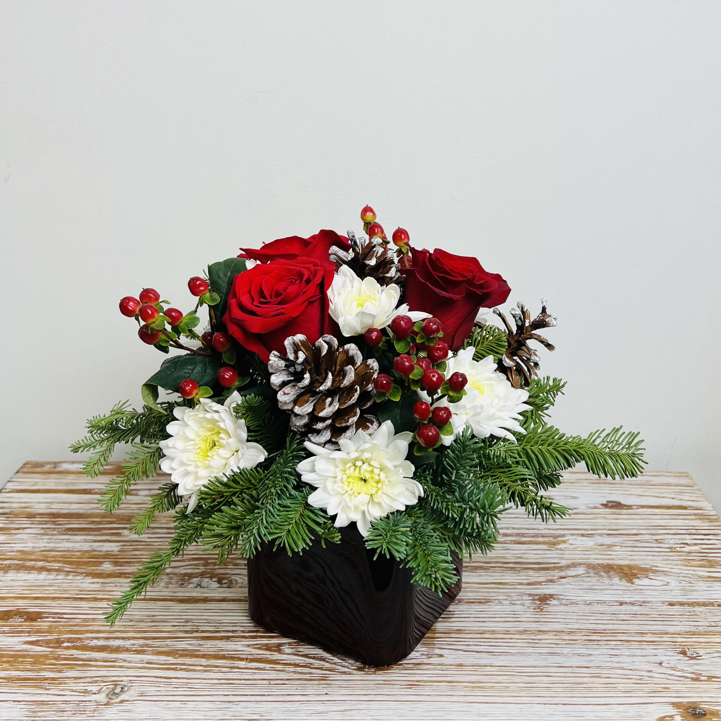 Berry and Bright - A wonderful natural winter arrangement of red roses and white cushion poms accented with red berries, white-tipped pinecones, and winter greens that all come together in a red cube vase.  Approximately 10” Tall