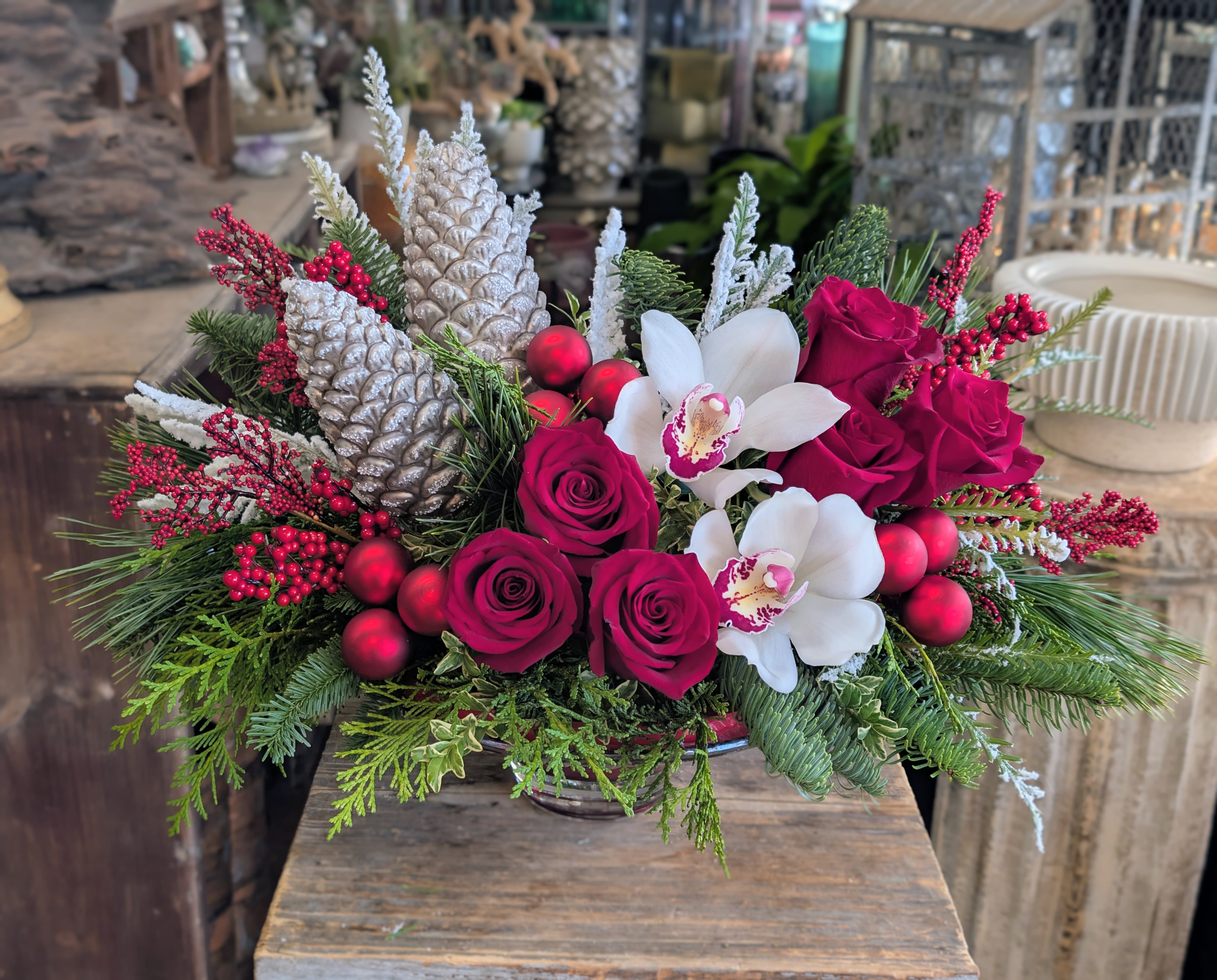 Marguerite - Marguerite is a graceful nod to classic French winter floristry, blending old-world elegance with festive seasonal warmth. Velvety crimson roses unfold alongside pristine white cymbidium orchids, creating a striking yet refined contrast reminiscent of European holiday tables and candlelit winter evenings. Lush evergreen foliage forms a textured foundation, accented with frosted glass pinecones, winter berries, and subtle holiday elements that evoke timeless seasonal charm.  Designed in a low, artful silhouette, Marguerite is ideal as a holiday centerpiece, a sophisticated seasonal gift, or an elevated statement for winter entertaining. Every detail is thoughtfully composed to feel nostalgic yet modern—rooted in tradition while remaining effortlessly luxurious.  Handcrafted by Windmill Floral Expressions in Las Vegas, Marguerite reflects our signature approach to custom holiday florals: intentional design, premium blooms, and enduring beauty. Perfect for Christmas celebrations, winter gatherings, or refined seasonal décor, this arrangement brings warmth, elegance, and classic French influence into any space.  Please note, this is a one sided arrangement. If the intention is to use this arrangement for a table centerpiece to be viewed from both sides, the premium size will accommodate this.  Available for local delivery in Las Vegas and Henderson. Custom color palettes and seasonal enhancements available upon request.