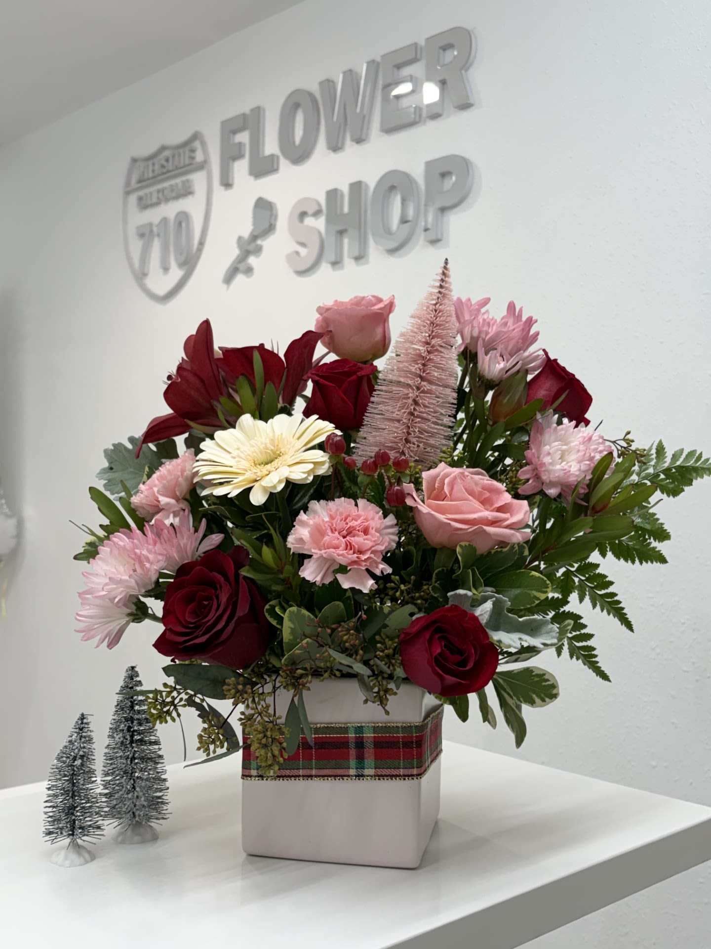 Blushing Holiday Charm - Soft, romantic, and festive, Blushing Holiday Charm. Designed in a chic keepsake container with a plaid ribbon accent, this arrangement balances holiday tradition with a fresh, elegant palette. Ideal for Christmas gifting, winter birthdays, or anyone who loves a softer take on seasonal florals. (If some flowers and/or the vase are not available there will be minor substitutions.)