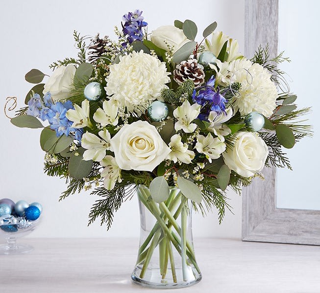 Season’s Wishes Bouquet - Send wonderful winters wishes with our luxurious arrangement. Snowy white blooms are gathered with lush greenery, accents of blue, gray and glistening silver &amp; gold. Loosely gathered in a glass vase, it’s a gift designed with stunning seasonal style.