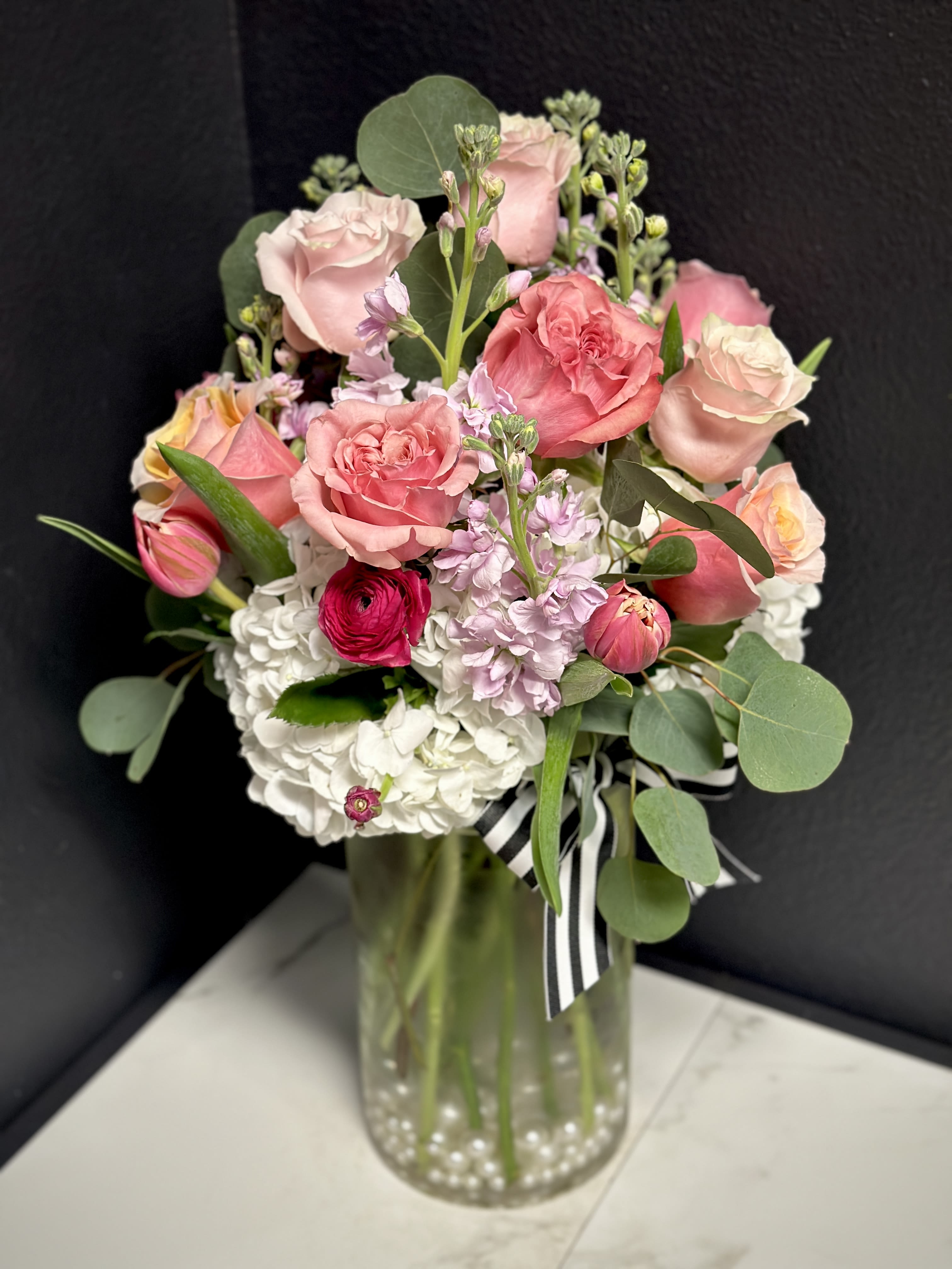 Pink Magpie Signature Rose Garden - Our Magpie Signature Rose Gardens are sure to impress. One or two dozen mixed pink roses are accented with hydrangea &amp; seasonal blossoms. Arranged in a clear glass vase with pearls. Rose colors will vary and may include shades of soft to hot pink. Pictured as Standard - 1 Dozen Roses. Deluxe - 18 Roses Premium- 2 Dozen Roses Approximately 17" tall. 