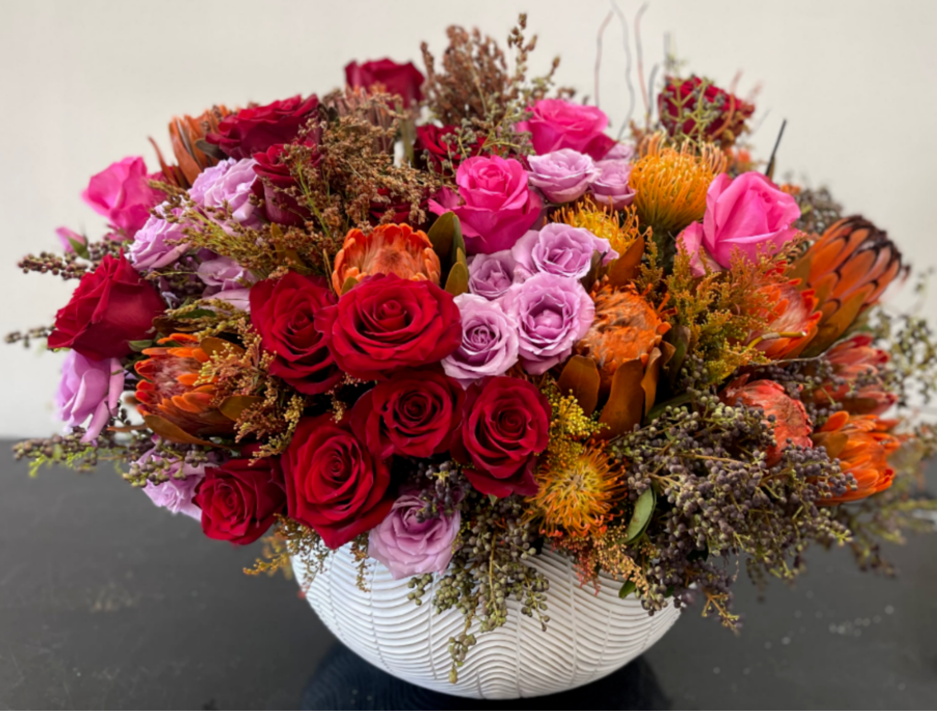 Holiday Jewel Rose Centerpiece - A bold and festive holiday centerpiece featuring rich red roses, lavender accents, vibrant protea, and seasonal textures. Designed low and full, this arrangement offers dramatic color and modern holiday elegance, perfect for statement tables and elevated gifting.  Hand-designed with premium fresh cut flowers and delivered fully arranged in a decorative vessel.   Note: Exact flowers may vary slightly based on seasonal availability while maintaining overall color palette, size, and style.