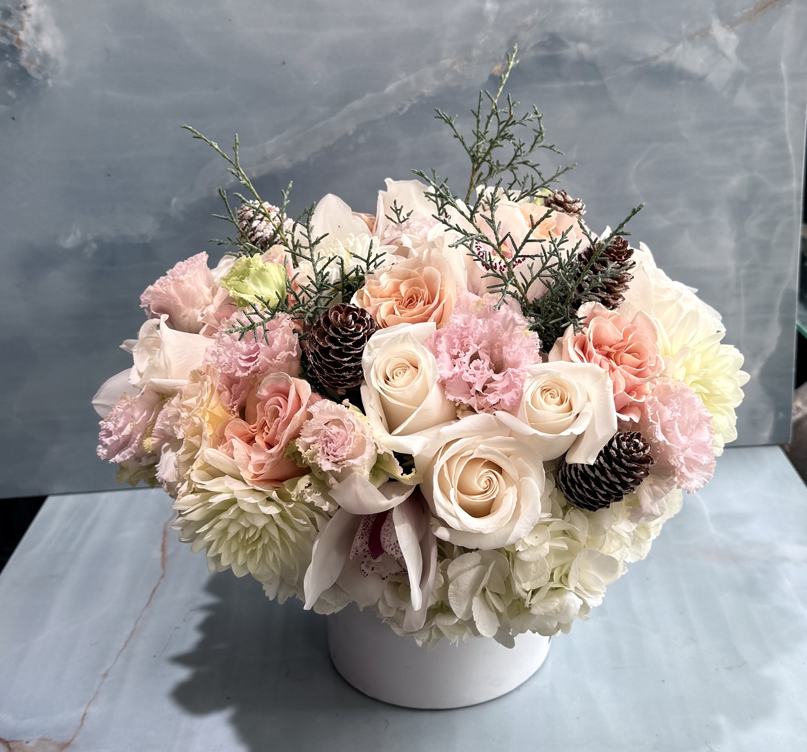 Blushing Winter Rose - This very soft, romantic, and luxurious arrangement is designed in a low, white ceramic vessel with lush, rounded silhouette that feels full yet airy. Creamy ivory and blush roses take center stage, with pink lisianthus and white hydrangeas. This arrangement is perfect for winter celebrations, elegant holidays, or a gentle, romantic statement centerpiece. 