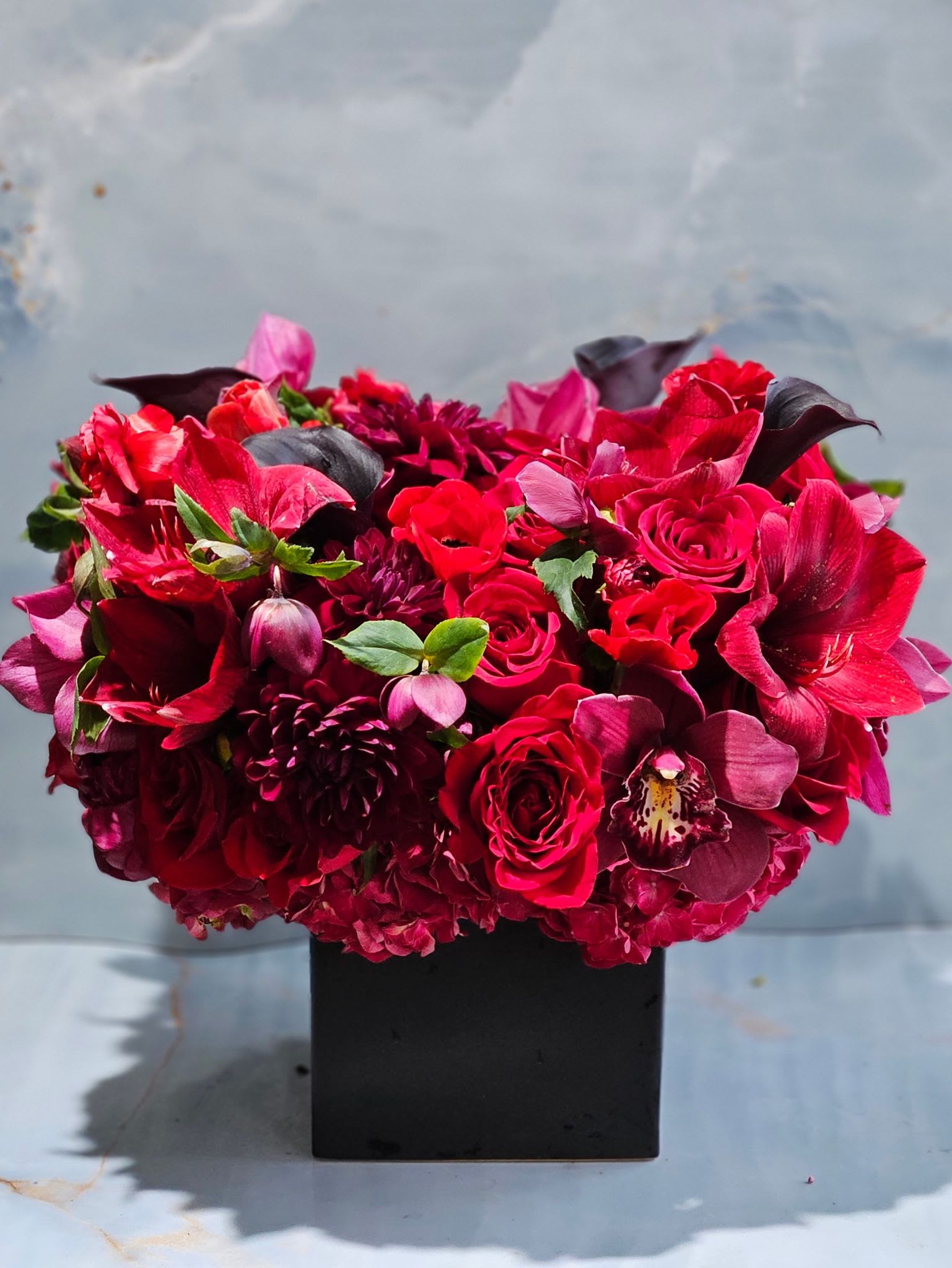 Ruby Opulence  - This striking floral arrangement is a lush, modern composition designed to make a bold statement. Featuring rich tones of crimson, burgundy, and deep wine, it blends velvety roses, sculptural calla lilies, and layered seasonal blooms. Set in a sleek matte black cube vase, the arrangement feels contemporary and luxurious, with strong color saturation and elegant texture throughout. 