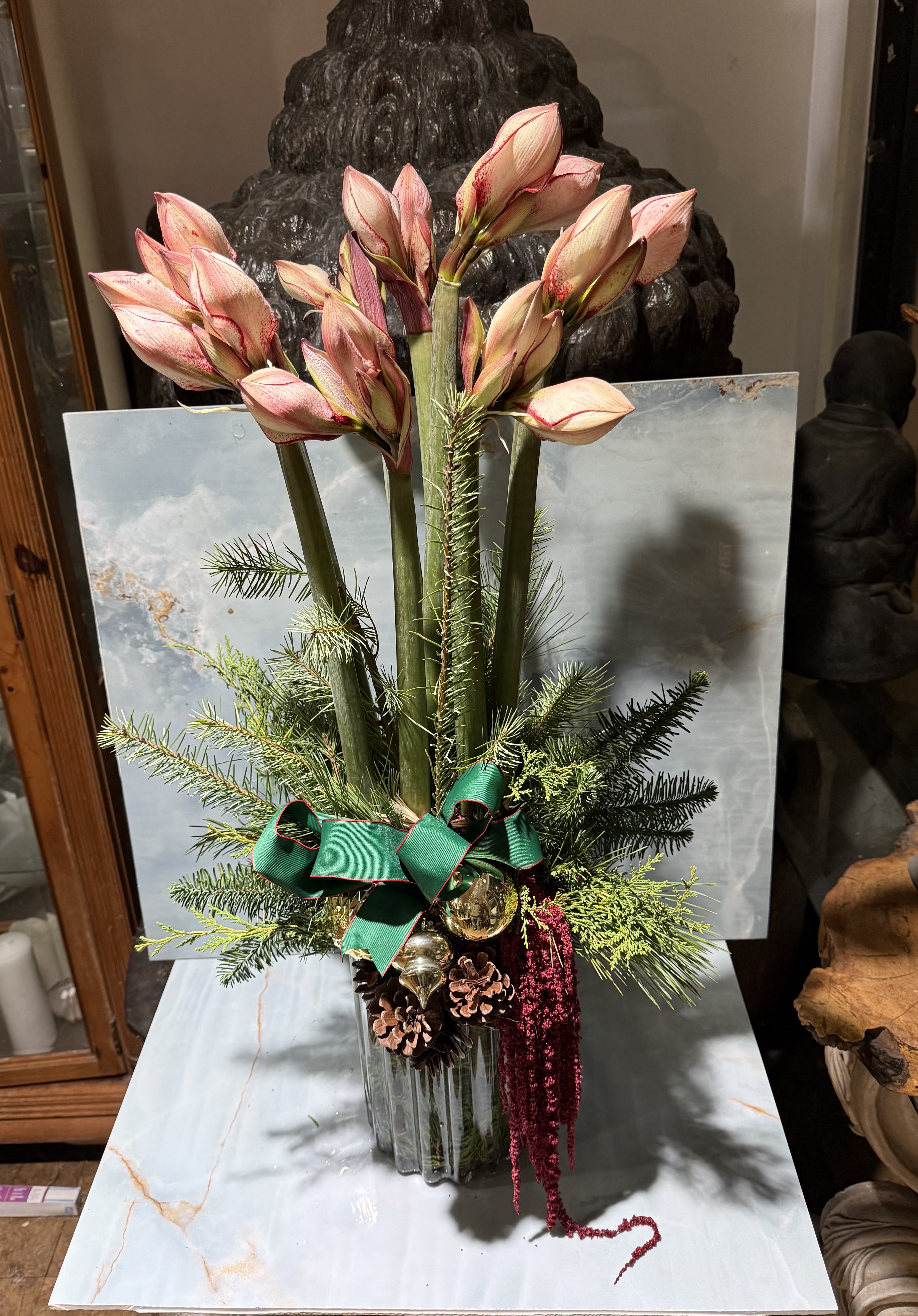 Holiday Amaryllis  - This arrangement is dramatic, elegant, and seasonally refined, with a strong architectural presence. Tall blushing amaryllis buds rise gracefully from the center, creating height and a sense of anticipation as they prepare to open. At the base, natural pinecones, gold ornaments and cascading amaranthus introduce warmth, richness, and movement.  