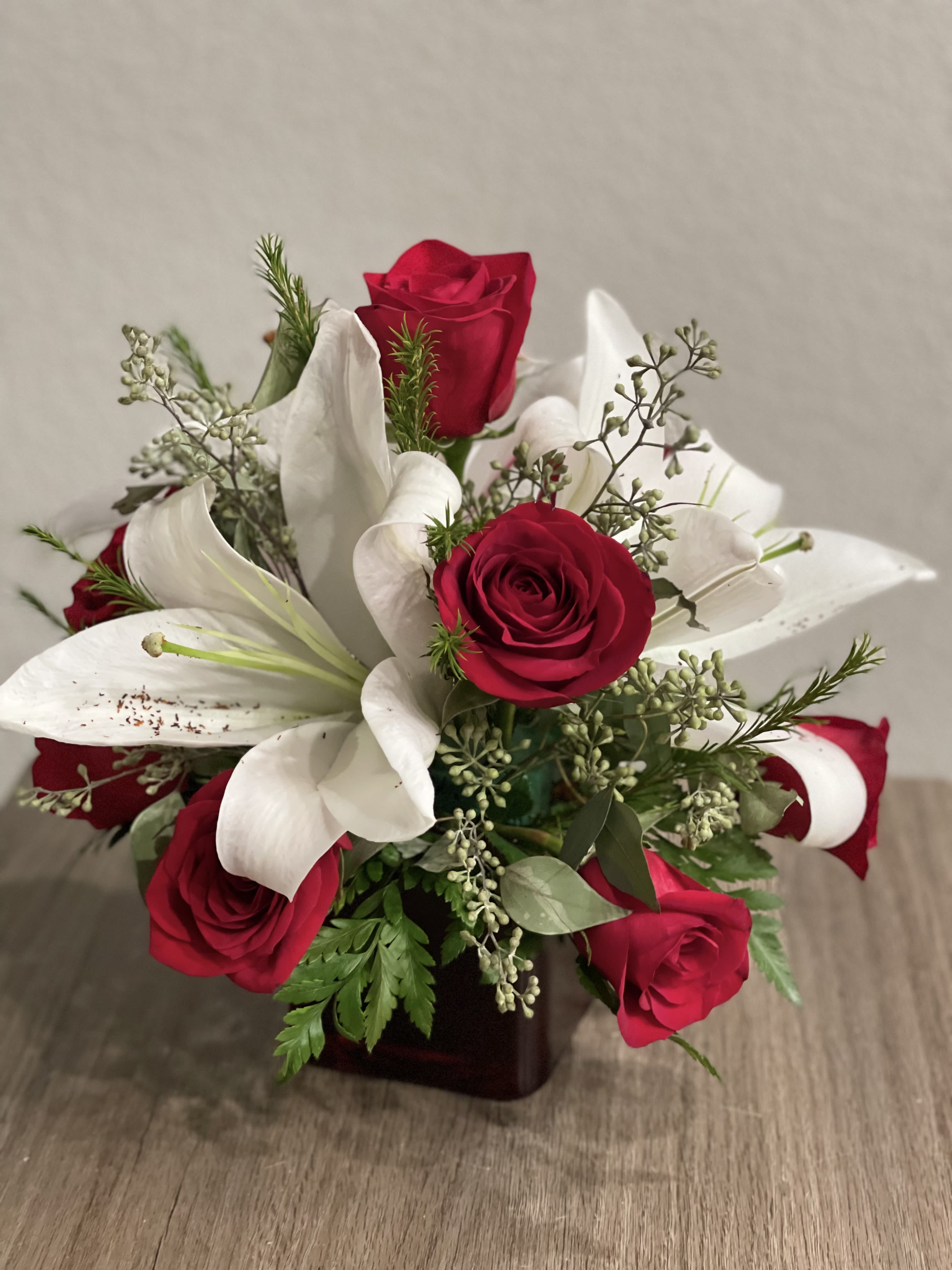 Budding Gifts - Cheerful floral arrangement of  red premium roses, white Asiatic lilies and assorted greenery all around on a square glass red vase.