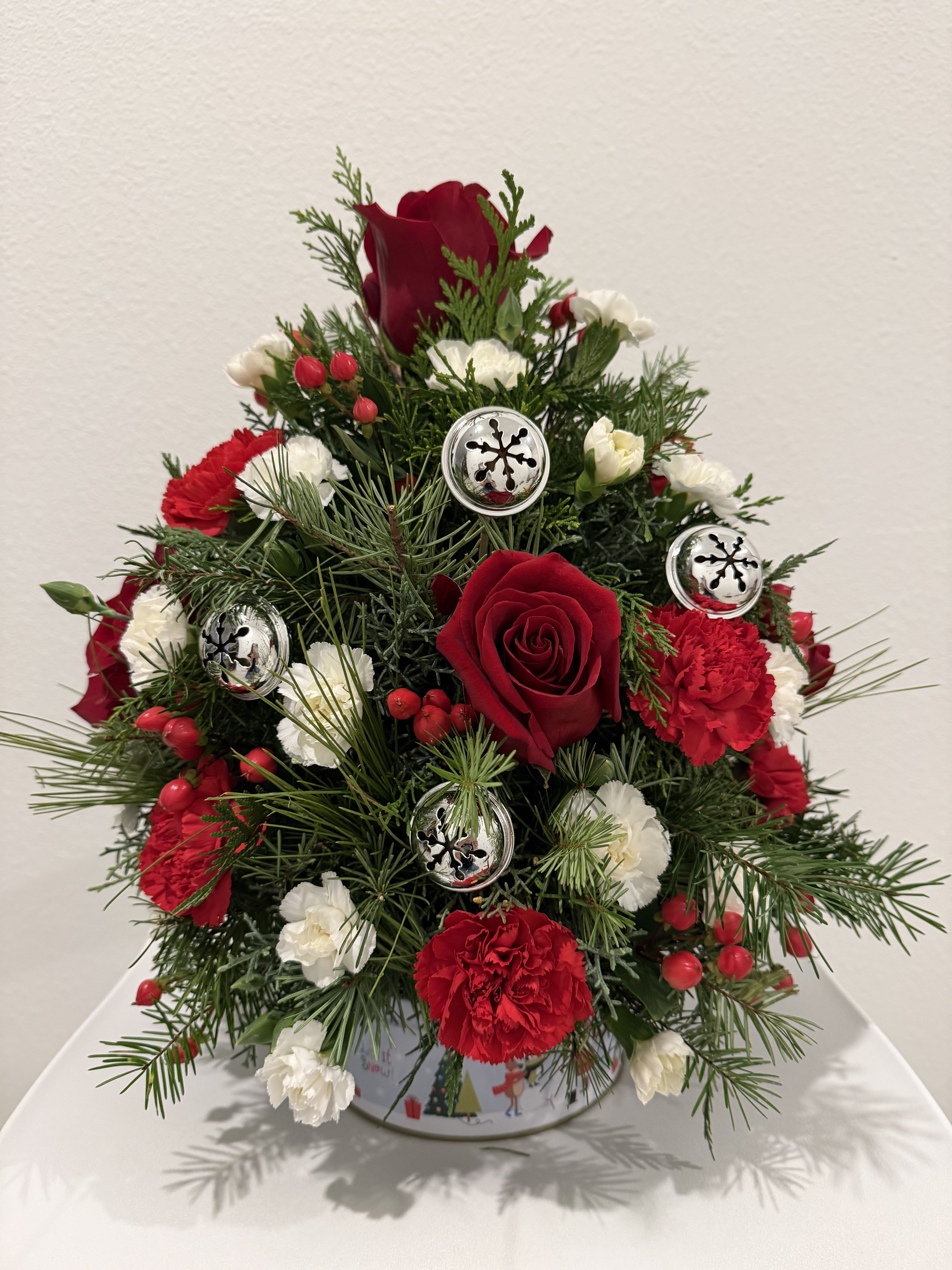 Christmas Tree of Roses - A festive Christmas Tree–style floral arrangement designed with rich red roses and red carnations, complemented by delicate white mini carnations. Lush evergreen pine and seasonal greenery create a classic holiday look, while bright red berries and silver jingle bell ornaments add a joyful Christmas touch. Arranged in a charming holiday container, this piece brings warmth, tradition, and seasonal elegance to any home or celebration. 