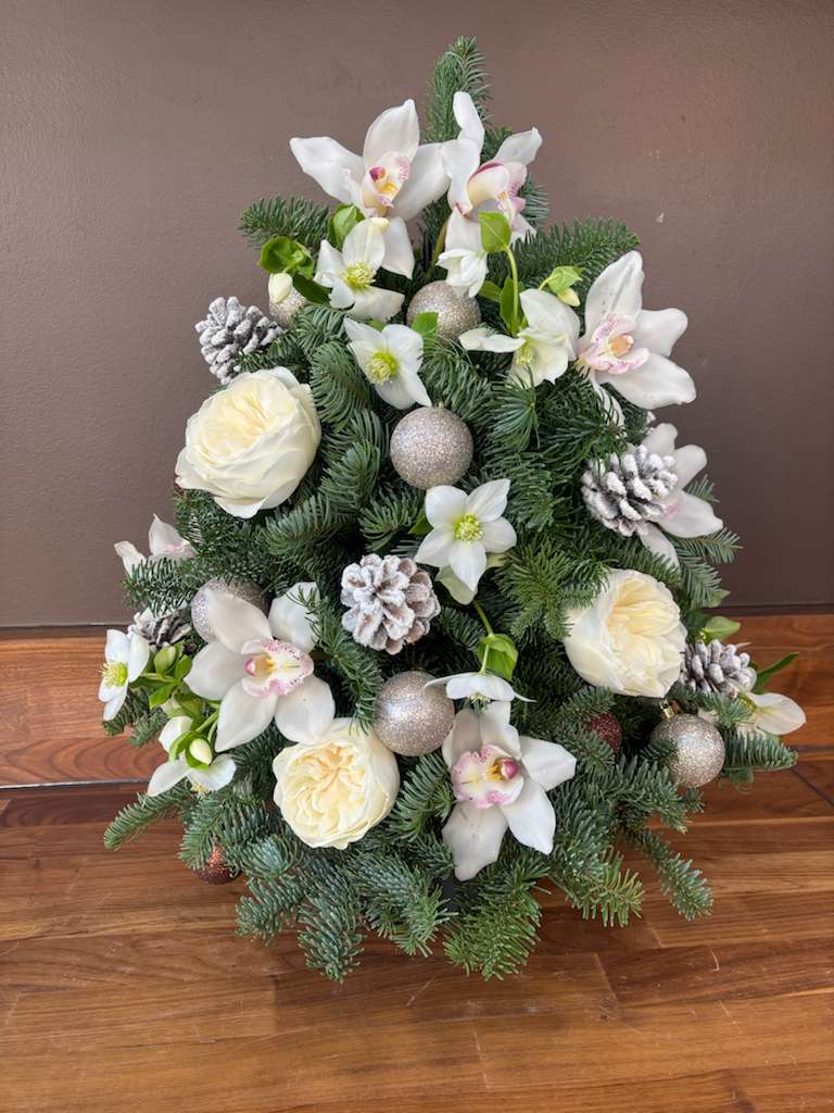 Holiday Cheer - This Holiday Cheer arrangement is shaped like a miniature Christmas tree, crafted from lush pine branches and adorned with snowy white roses and delicate white orchids. Accents of white-painted pinecones and glittering silver ornaments add a festive sparkle, creating a wintry, elegant centerpiece perfect for seasonal décor. The combination of natural greenery and refined white florals evokes both celebration and serenity, making it ideal for gifting or display during the holidays.