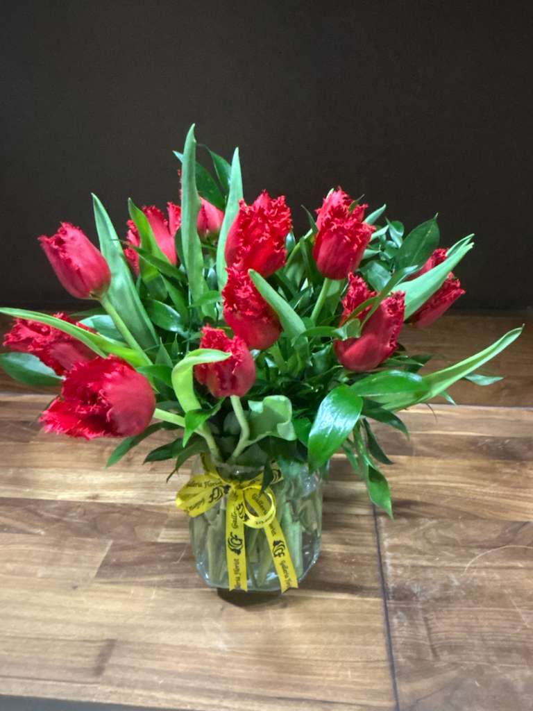 Very Tulips - This vibrant arrangement showcases a cheerful burst of red tulips with fringed petals, surrounded by lush green leaves and tied with a decorative yellow ribbon printed with black text. Presented in a clear glass vase, the bouquet feels fresh, modern, and full of personality—perfect for gifting or brightening up a space with bold simplicity.