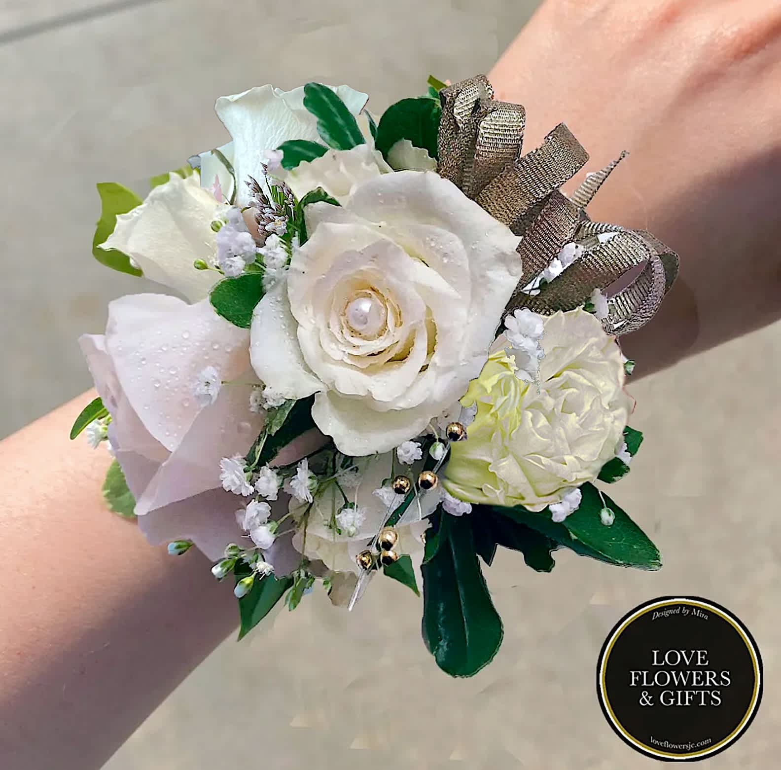 Wrist corsage of white and pale blush roses with baby’s breath and gold ribbon on a person’s wrist.