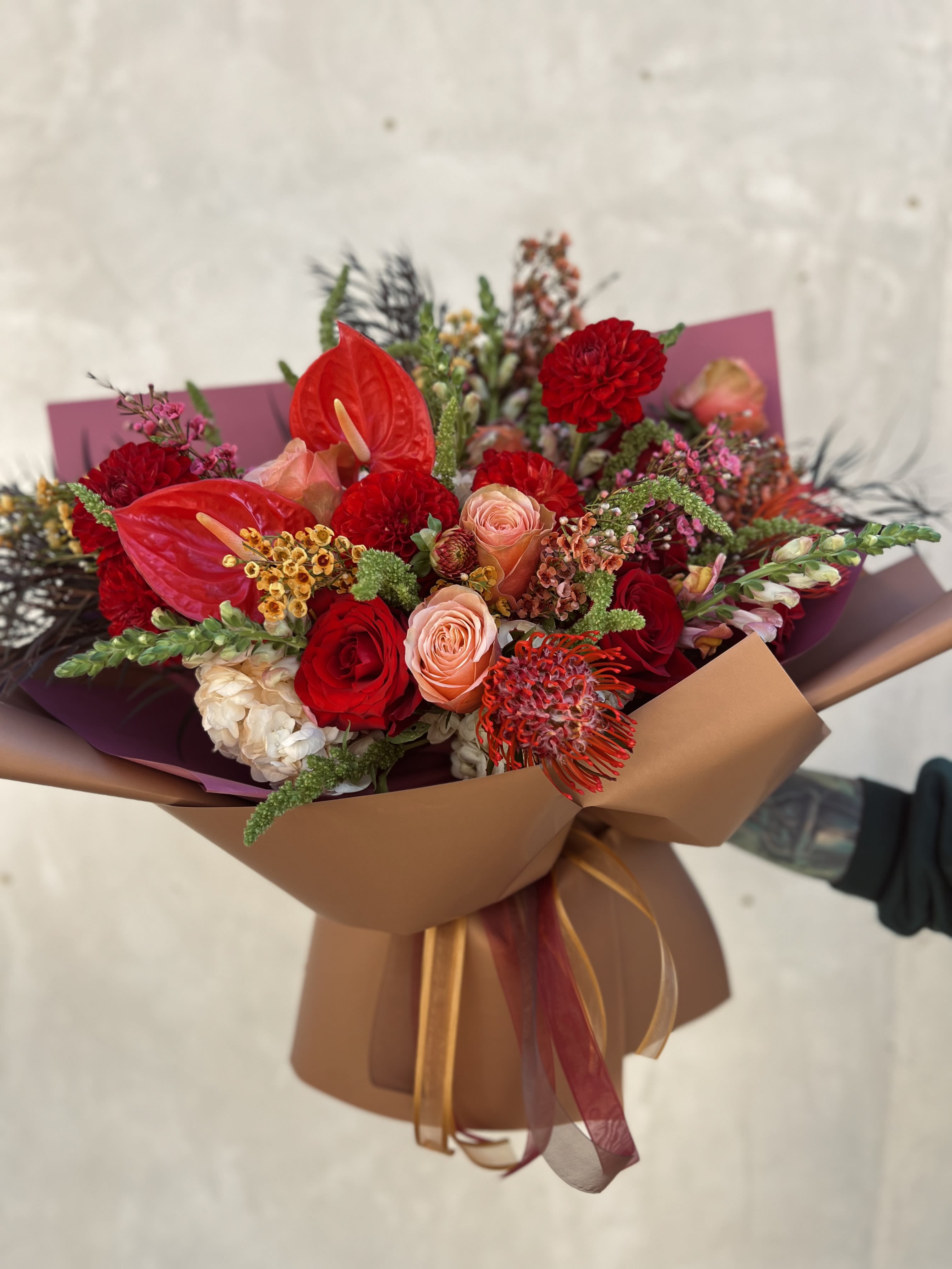 Gingerbread Bouquet  - Flowers: kahala roses, freedom roses, pin cushions, red anthuriums, red dahlias, upright green amaranthus, orange hydrangeas, burgundy willow eucalyptus, fall waxflower, and snapdragons.   Wrapped in red/copper paper with a red and copper ribbon. 