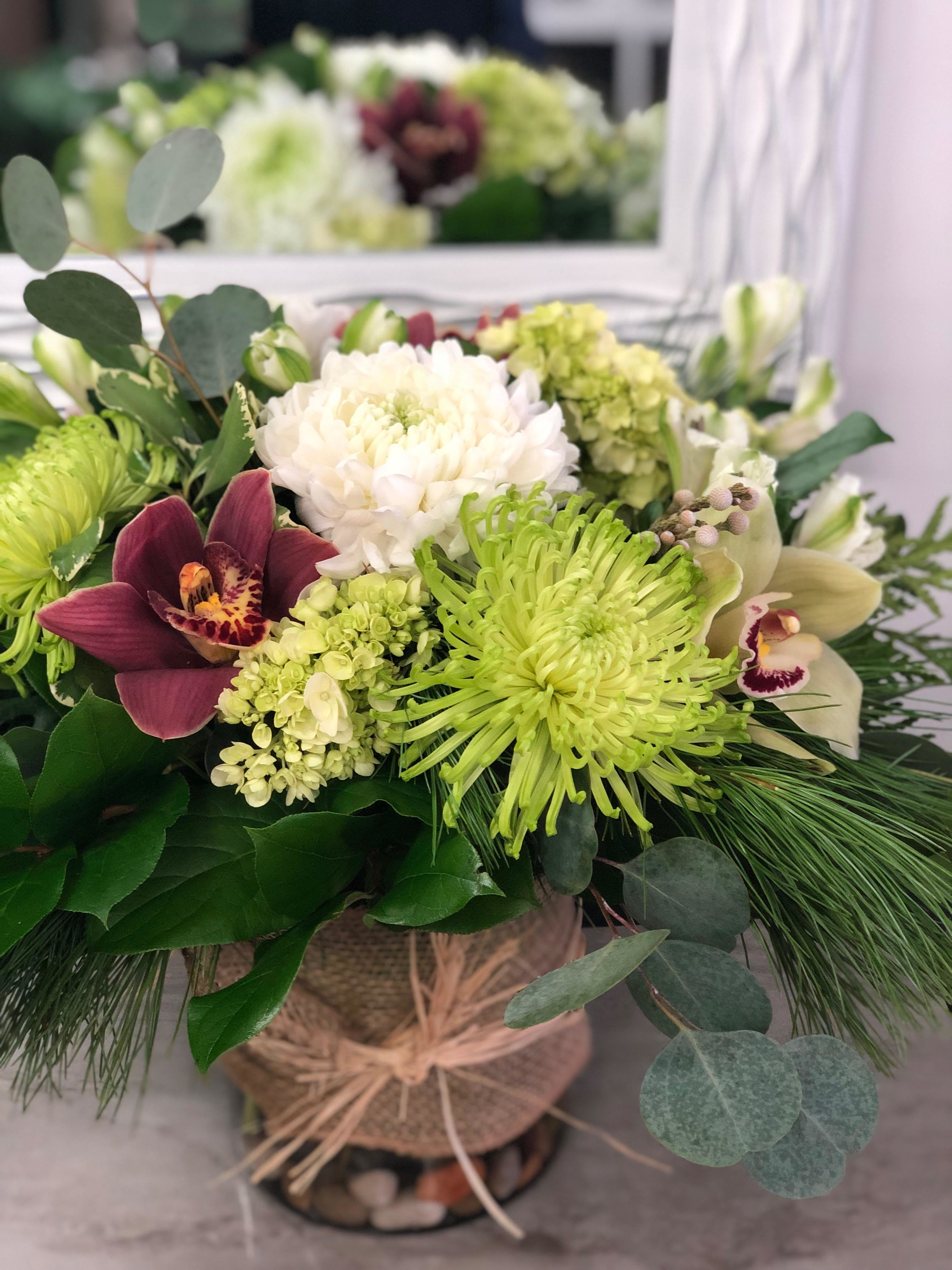 Burlap &amp; Burgundy - A rustic Christmas arrangement featuring crisp white blooms, lush evergreens, and rich burgundy accents. Displayed in a burlap-wrapped vase, it blends cozy farmhouse charm with classic holiday colors.
