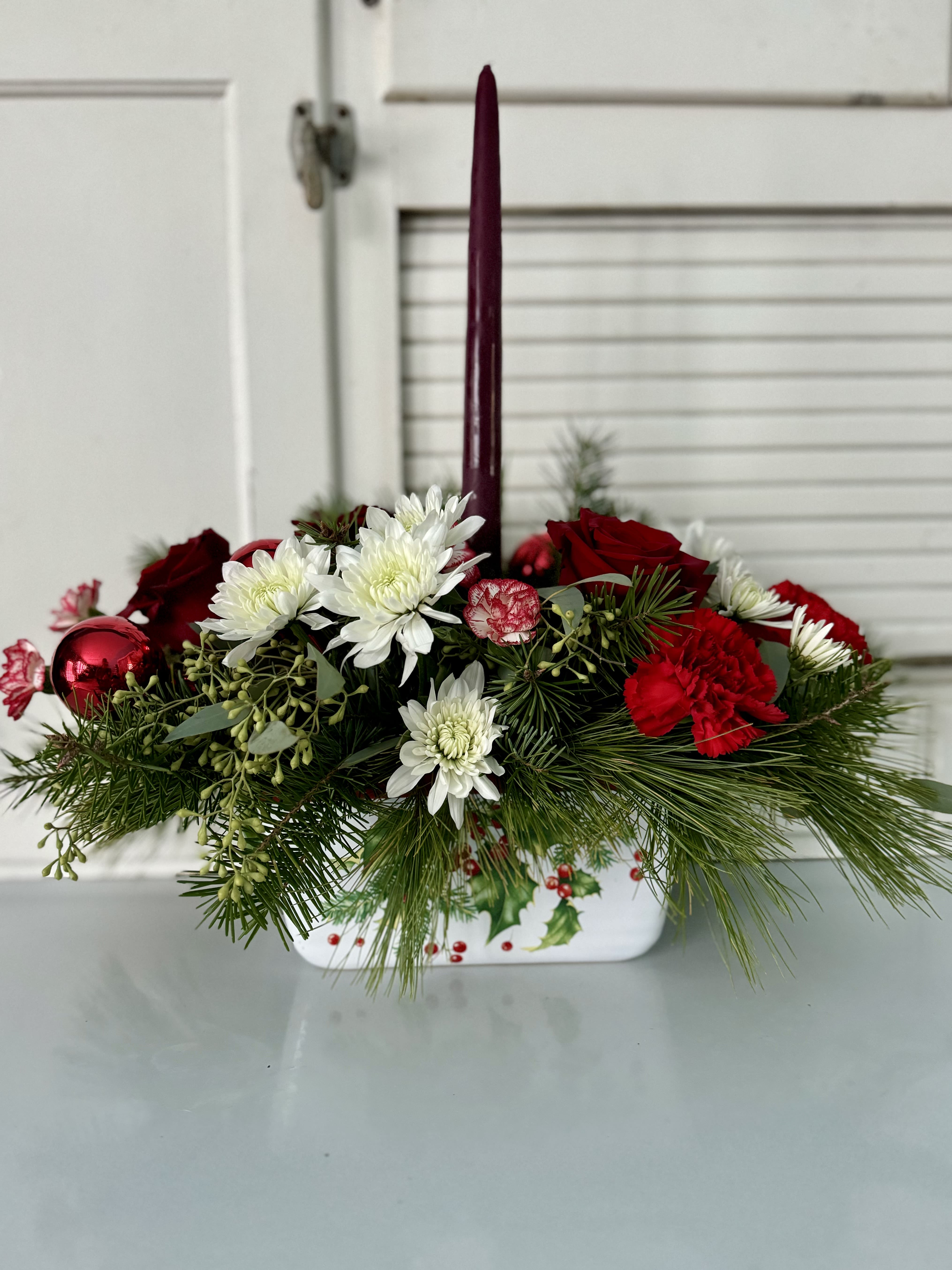 Holly Berry Elegance - A timeless holiday arrangement featuring red carnations, white chrysanthemums, and festive greenery, nestled in a holly-printed container. The central candle brings a cozy glow to the heart of your table.