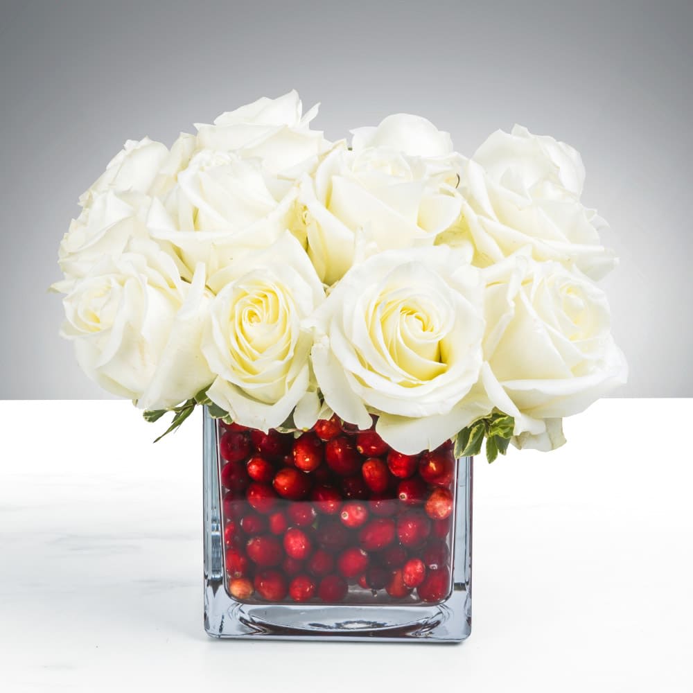 The Cranberry Rose - You appreciate design that speaks through contrast. In a season of "more is more," there is profound elegance in clean lines and striking color.  The challenge: finding holiday decor that feels crisp and modern. You want an arrangement that feels less like a bouquet, more like an art piece.  This is The Cranberry Rose. A study in chic opposites, pairing twelve snowy white roses with the festive pop of crimson cranberries. Housed in clear glass, the berries create a vibrant, jewel-toned base anchoring the pristine blooms.  Architectural and clever. Placed on a cocktail table or dining console, it captures the light and draws the eye. The perfect blend of winter frost and festive fruit. A modern classic for the Naples home. Every bloom tells a story.  Perfect for: Naples modern holiday decor, architectural floral arrangements, elegant hostess gifts, and sophisticated red and white centerpieces.  How do I style a modern Christmas cocktail table? Keep it focused. Instead of scattering trinkets, choose one bold, architectural statement like this. The contrast of white roses and red berries provides all the festive color needed without cluttering the surface where guests set their drinks.  We are now accepting reservations for your holiday gatherings in Naples, Marco Island, and Bonita Springs. We strongly encourage securing your delivery date now, as our calendar fills quickly.