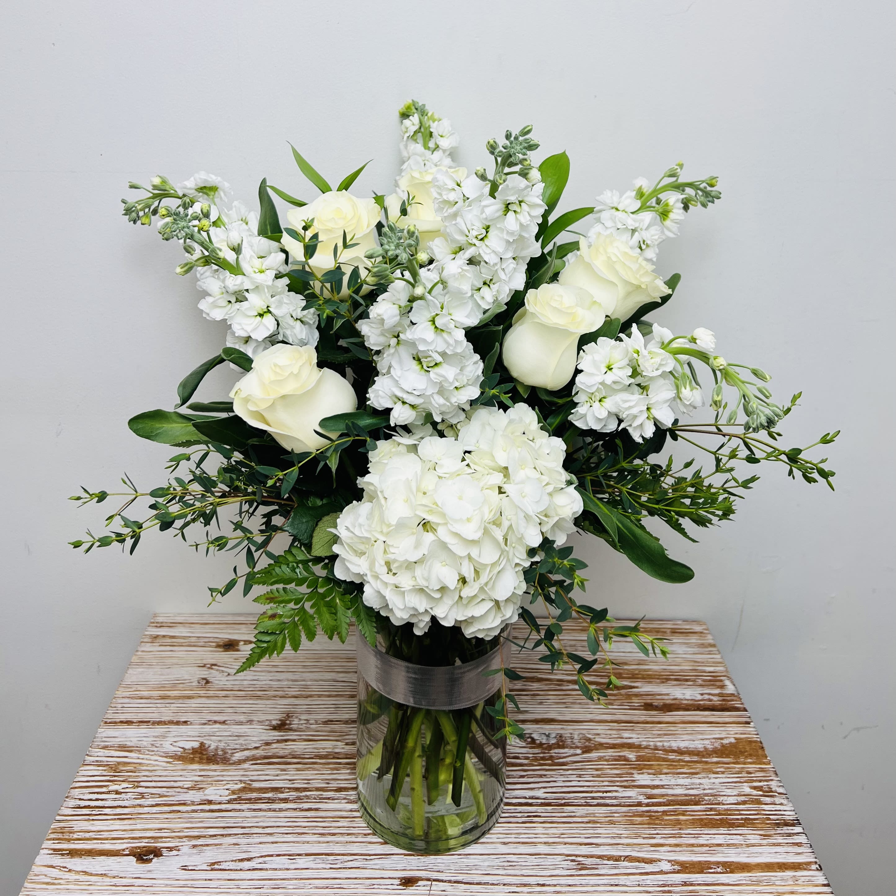 Sincere Sympathy - A perfectly fitting way to show your sympathy for a loss of a loved one.  This arrangement contains blooms of white, including Hydrangea, Roses, Stock and other seasonal blooms in a clear cylinder vase accented with silver ribbon.  Approximate Dimensions: 16” Tall