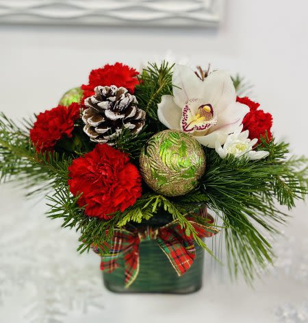Holiday Blooms - Celebrate the season with a charming holiday centerpiece featuring vibrant red carnations paired with an elegant white orchid bloom. Nestled among lush fresh winter greens—such as cedar, pine, and fir—this arrangement brings the warm, fragrant spirit of Christmas indoors. The flowers are artfully designed in a modern square glass vase, wrapped with festive tartan ribbon for a classic holiday touch. Perfect for gifting or adding a cozy, traditional accent to your home décor.