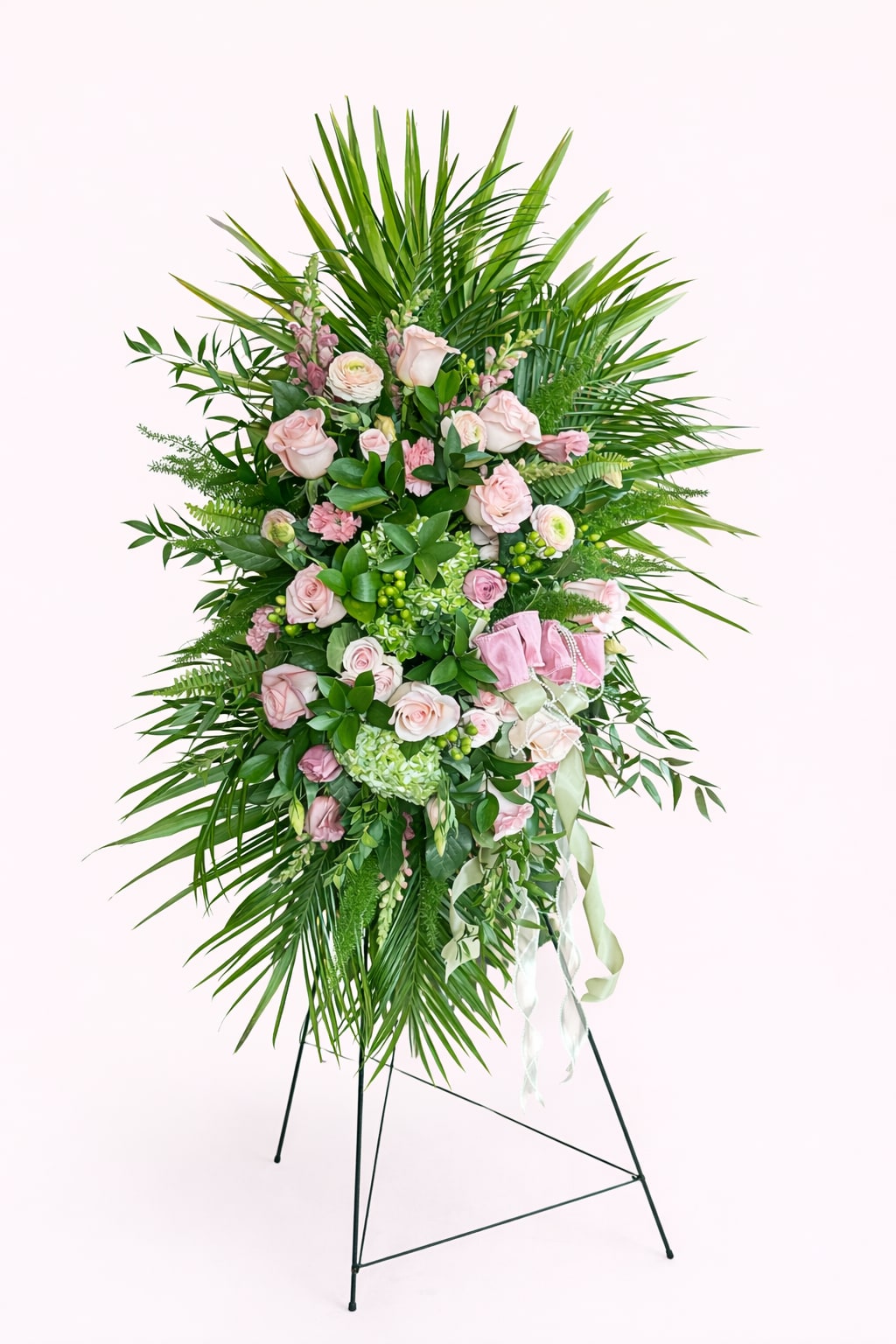 Blush Serenity Standing Spray - The Blush Serenity Standing Spray is a graceful sympathy arrangement designed to honor life and express heartfelt condolences. Featuring soft blush roses, lush hydrangea, delicate pink accent blooms, and abundant greenery with flowing palm fronds, this standing spray creates a full, elegant presentation suitable for funeral services, memorials, and celebration-of-life ceremonies.  Expertly handcrafted by our florists, this pink and green standing spray offers a serene and comforting presence while conveying love, remembrance, and sympathy. Displayed on an easel, it serves as a beautiful tribute for services held at funeral homes, churches, or graveside ceremonies.  ⸻  Details: 	•	Arrangement style: Standing spray on easel 	•	Color palette: Blush pink, soft green, and fresh greenery 	•	Occasion: Sympathy, funeral, memorial, celebration of life 	•	Designed and delivered by Bloom-A-Round Floral Design