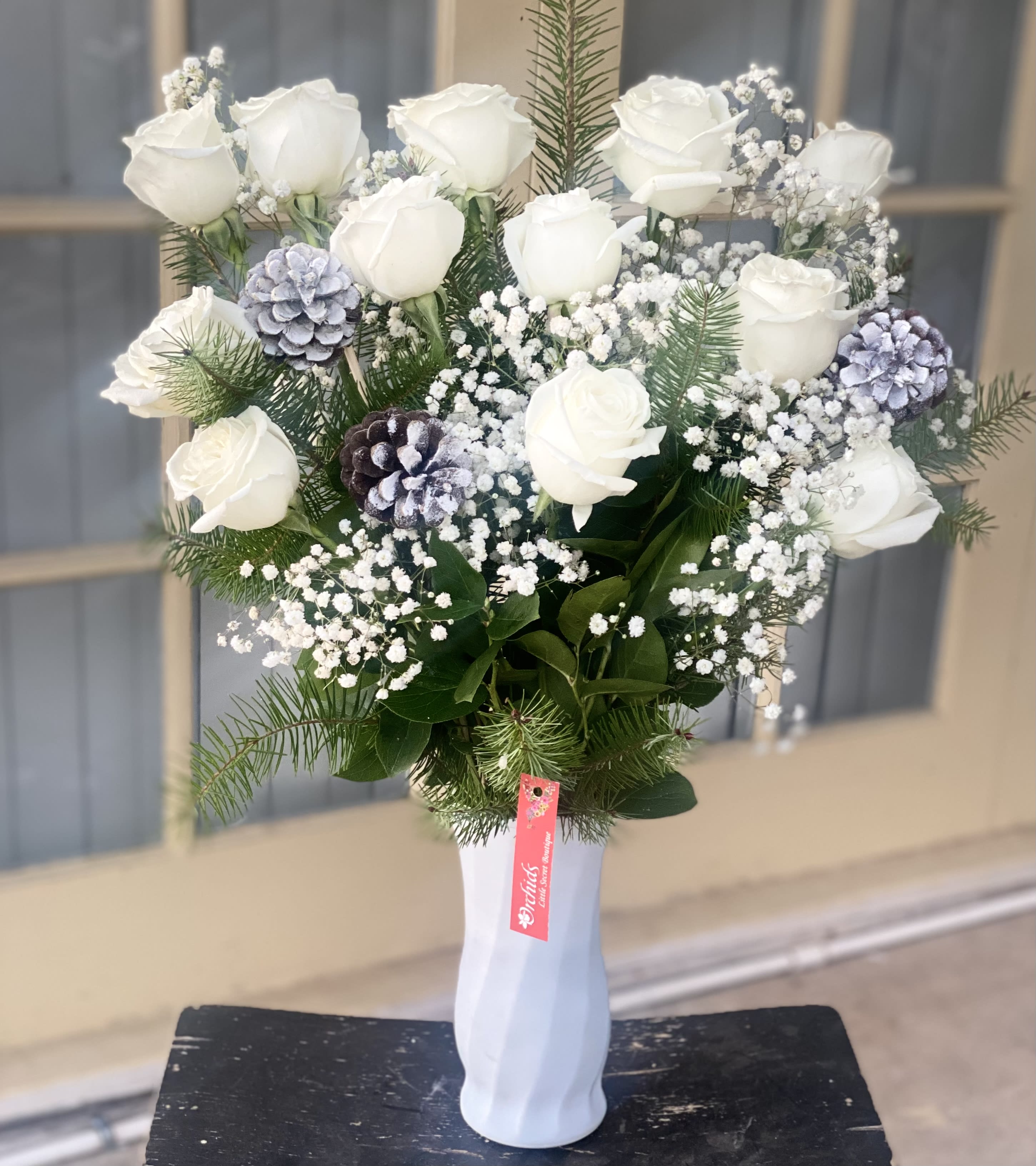 Snowy White Roses &amp; Pinecones  Bouquet  - Fresh as  fallen snow! Snowy white premium roses are decorated with winter pinecones and holiday greens arranged in a white vase. Perfect for any winter celebration, gift to offer congratulations for graduation, engagements, bridal showers, new baby, or even a gift of sympathy. Bouquet measures approximately 23”H  
