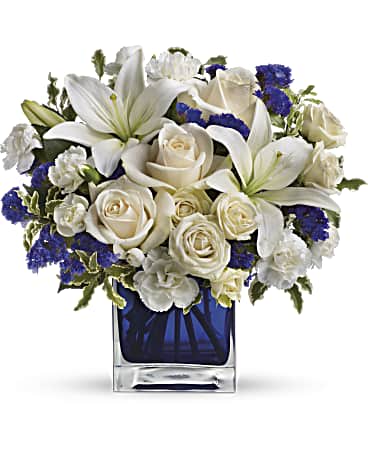 Sapphire Skies Bouquet - Send someone a bit of heaven with this beautiful bouquet. Luxurious creme roses and pure white lilies paint a peaceful picture inside a sapphire blue cube. Creme roses, white asiatic lilies and white miniature carnations are mixed with bursts of purple statice and green pitta negra. Delivered in a 4x4" glass Cube - arrangement measures approx. 11.5x11" for Standard size, 12.5x12" for Deluxe, and 13x13.5" for Premium.