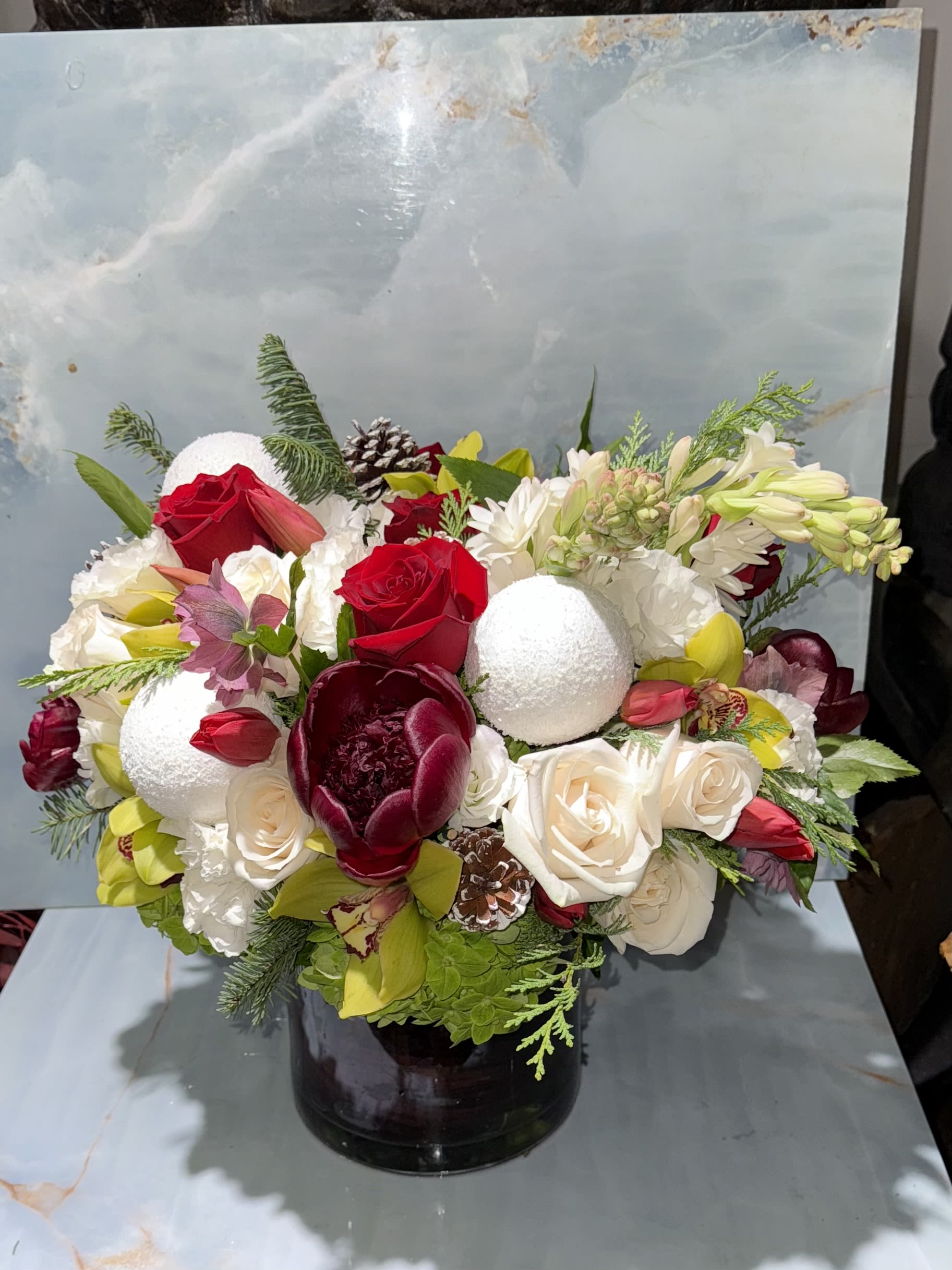 Fireside Elegance - This arrangement is a luxurious winter centerpiece that blends classic romance with seasonal charm. Rich crimson roses and burgundy blooms anchor the design, creating depth and warmth, white soft ivory and white flowers bring balance and elegance. Set in a sleek, dark vessel, the arrangement feels full, lush, and thoughtfully layered, with contrasting colors and textures that draw the eye from every angle. 