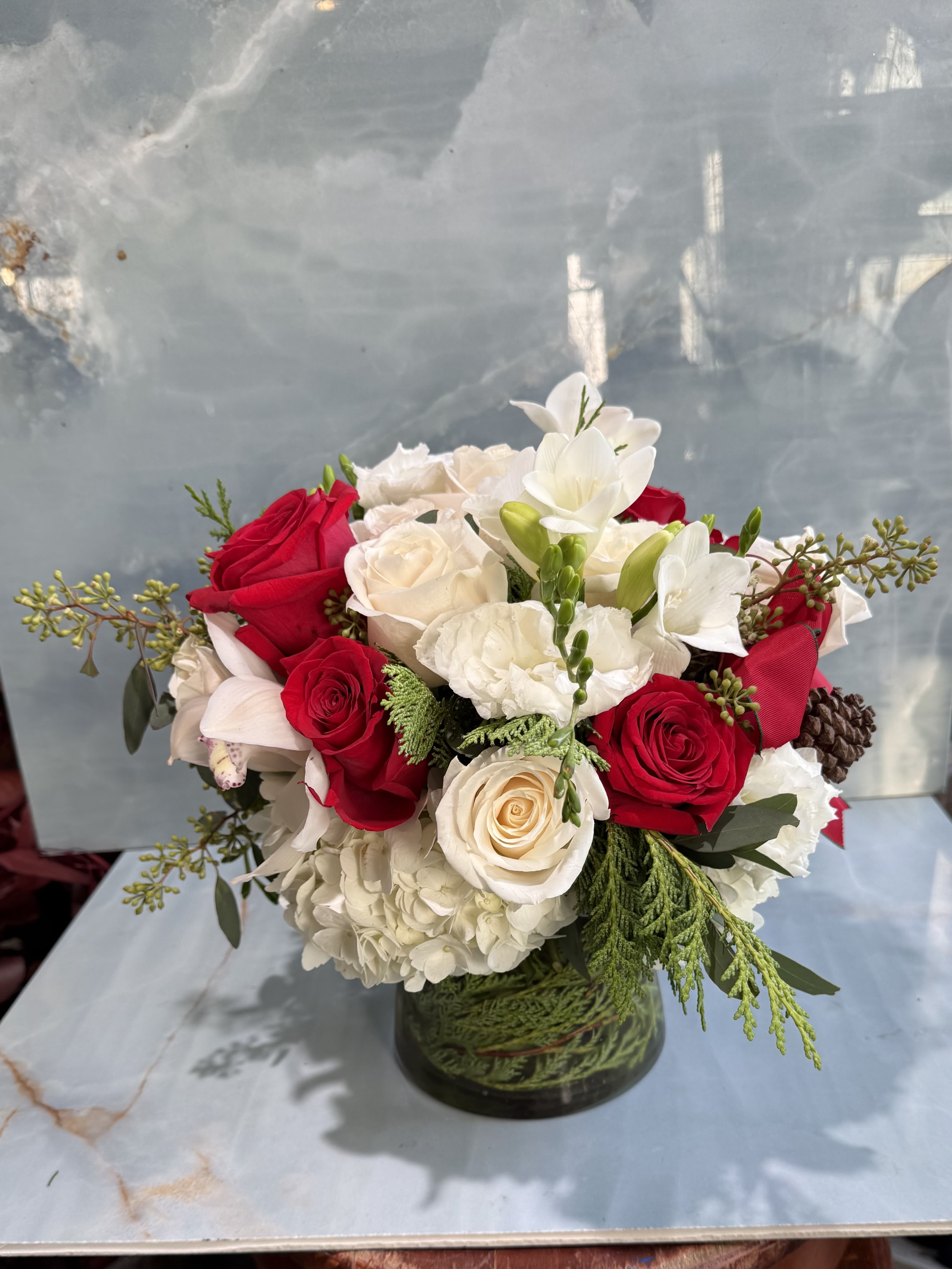 Scarlet &amp; Snow - This floral arrangement is elegant, classic, and warmly festive. A lush mix of velvety red roses and soft ivory blooms creates a timeless color palette that feels both romantic and refined. Creamy hydrangea forms a full, cloud-like base, while delicate white freesias and orchid accents add graceful movement and a light, airy touch. Designed in a clear glass cylinder with visible greenery below the waterline, the arrangement has a polished yet natural look. 