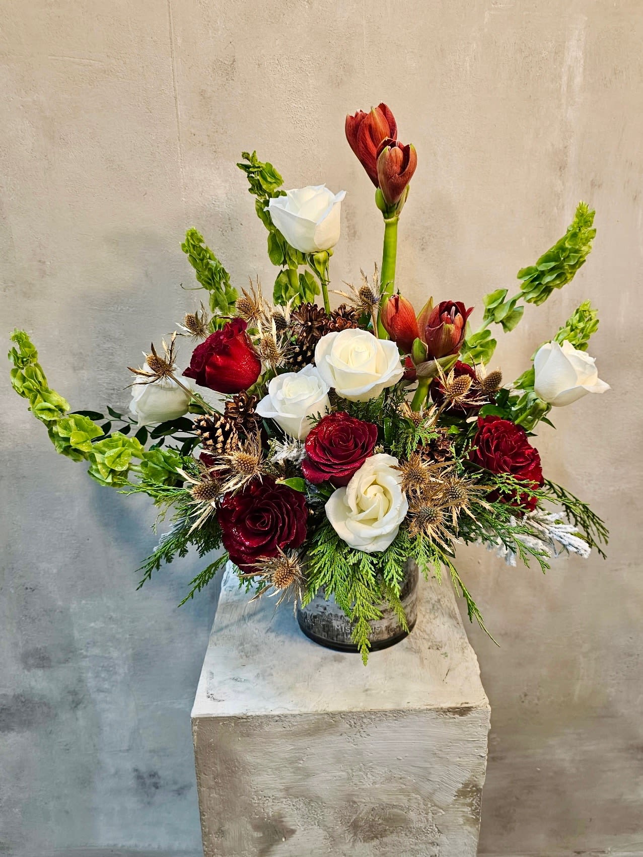 Grinch With A Twist  - This striking holiday arrangement blends timeless elegance with natural winter texture. Deep red and creamy white roses are artfully arranged with sculptural amaryllis, fresh seasonal greens, and rustic accents like pinecones and thistle for a warm, elevated feel. Designed in a clear glass vessel, this piece brings a refined yet cozy presence to any space.  Perfect for holiday gifting, winter celebrations, or as a statement centerpiece, this arrangement balances classic romance with modern, organic details. Each design is handcrafted using the freshest blooms, making every piece one of a kind.  Seasonal availability may vary; substitutions may be made while maintaining the overall color palette and style.