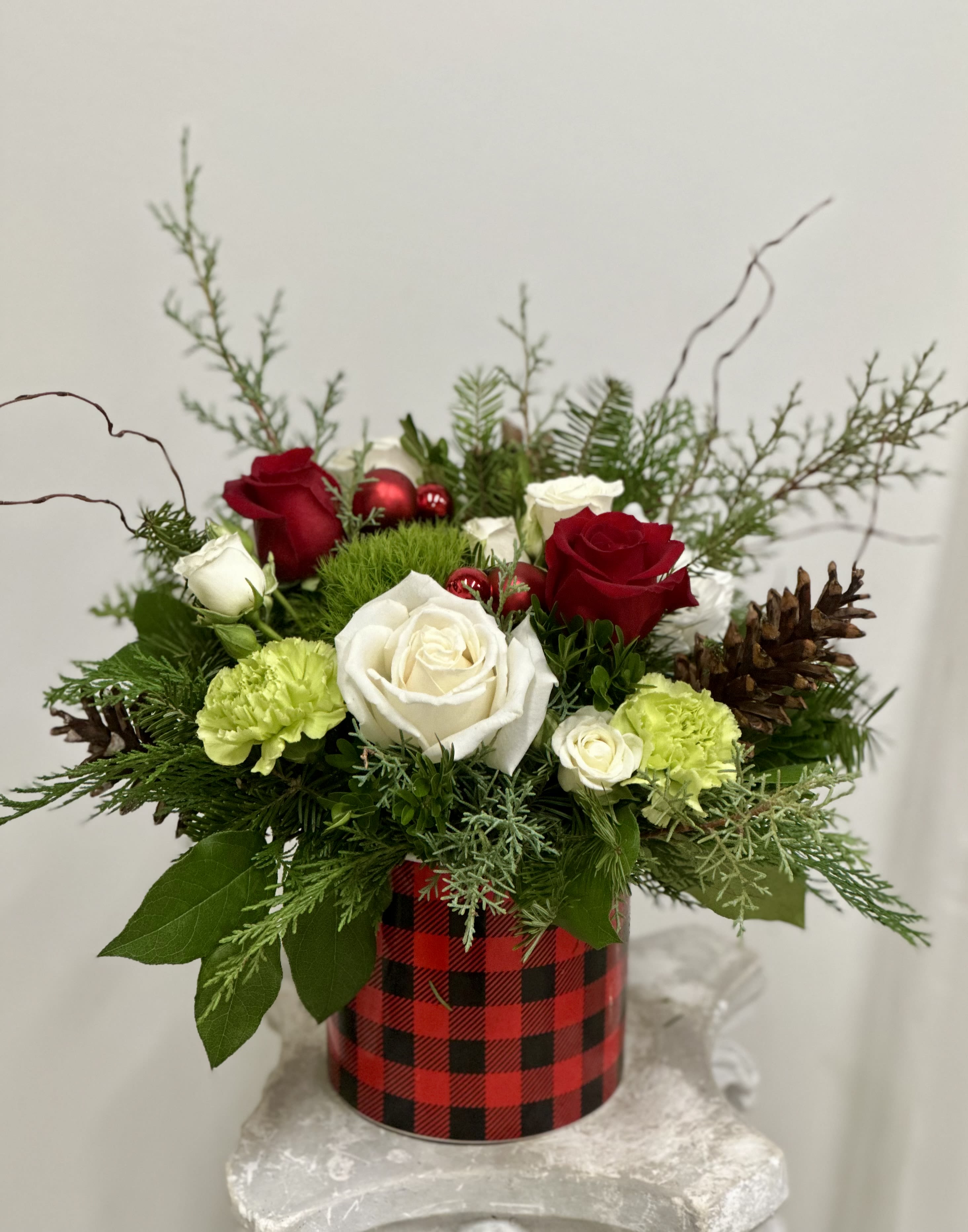 Cozy Christmas by Westford Florist Art by Veny - Embrace the spirit of the season with "Cozy Christmas" by Westford Florist Art by Veny, a limited-edition masterpiece that redefines holiday elegance. This enchanting arrangement features a harmonious blend of delicate white and red roses entwined with charming green trick, graceful white spray roses, and elegant white poms complemented by verdant green carnations. A touch of festive berries dances playfully among whimsical curly willow and delightful pinecones, while shiny ornaments add a splash of sophistication to this stunning display. Nestled within a stylish red and black plaid cylinder, “Cozy Christmas” is not just an arrangement—it's an invitation to indulge in the joy and warmth of the holidays. Transform your home into a winter wonderland and let every glance at this luxurious bouquet spark boundless cheer throughout your space. Perfect for gifting or as an opulent centerpiece for your own celebrations, it promises to be the highlight of any festive décor. Elevate your holiday experience; embrace cozy luxury today!