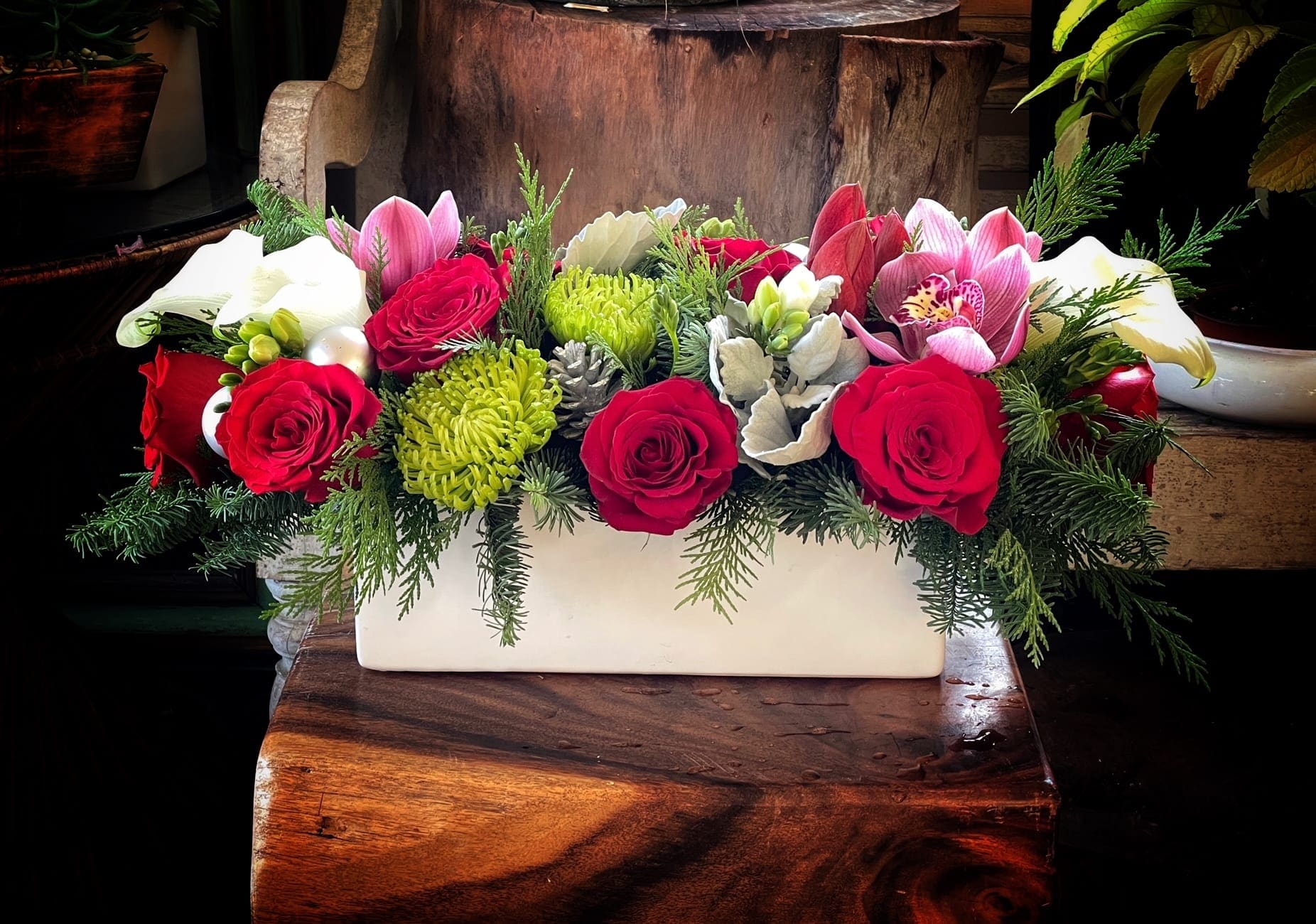 Santa Clause - A low, elongated centerpiece overflowing with velvety red rose, creamy white blooms (including calla lilies and ruffled whites), and deep burgundy accents, artfully nestled among seasonal evergreens. This arrangement evokes a sense of holiday abundance, timeless elegance, and cozy winter celebration-perfect for a Christmas dining table, mantel, or festive gathering. 