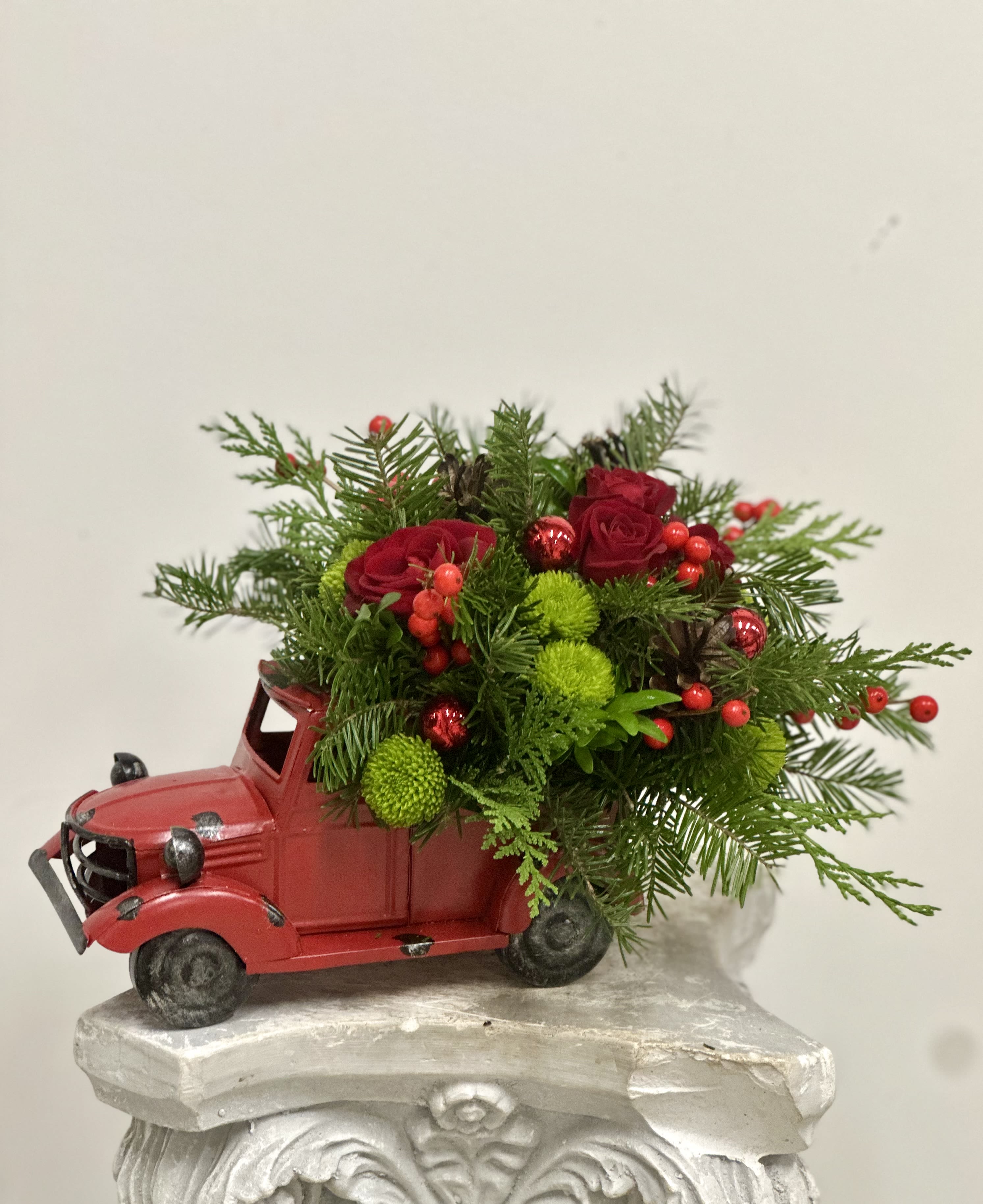 Merry and Bright Truck Arrangement by Westford Florist Art by Veny - Transform your holiday decor with the Merry and Bright Truck Arrangement by Westford Florist Art by Veny. This charming centerpiece features a vibrant red tin truck filled with a delightful mix of Christmas greens, luscious red roses, cheerful spray roses, and festive winter berries. Accentuated with eye-catching red glass ornaments, natural pinecones, and playful green dot poms, this arrangement captures the spirit of the season in every detail. Measuring approximately 11" by 8.5", it's perfectly sized to add a touch of whimsy to your table or mantelpiece. Elevate your holiday celebrations with this enchanting piece that effortlessly combines joy and elegance—an ideal gift for loved ones or a splendid treat for yourself. Celebrate in style!
