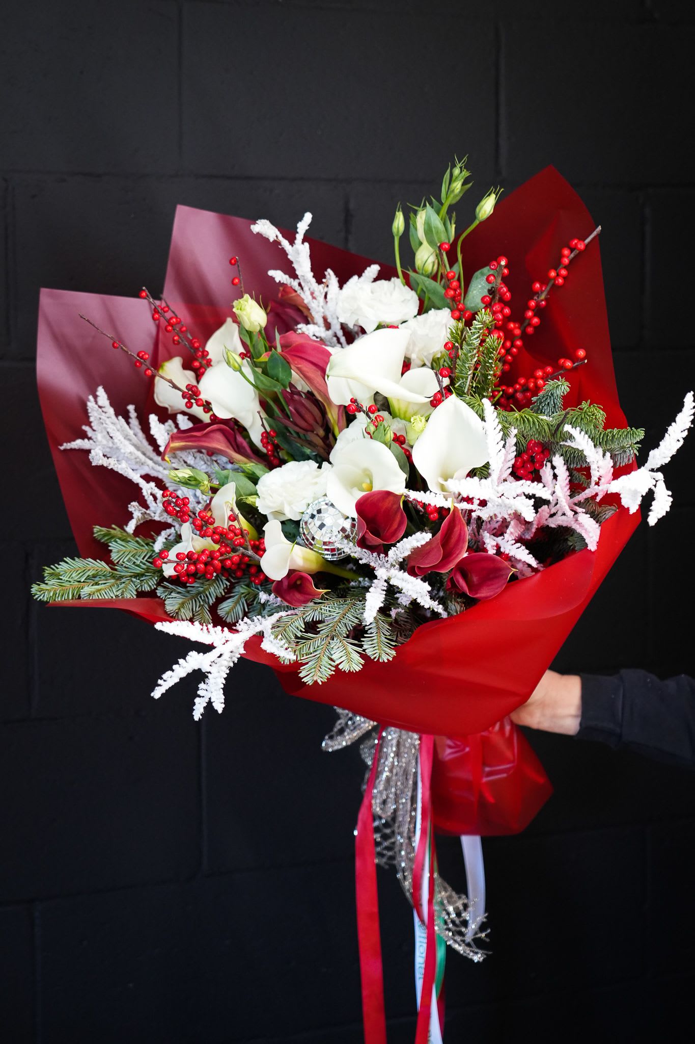 Crimson Frost Bouquet - The Crimson Frost Bouquet is a dazzling holiday arrangement that captures the festive spirit in every detail. Featuring deep red and white calla lilies, vibrant red berries, and delicate lisianthus, this bouquet is accented with frosted branches, evergreen sprigs, and glittering holiday elements. Wrapped in luxurious red paper and adorned with festive ribbons, it exudes seasonal cheer and elegance. Perfect for gifting or as a stunning centerpiece, this bouquet brings warmth and sophistication to any celebration.