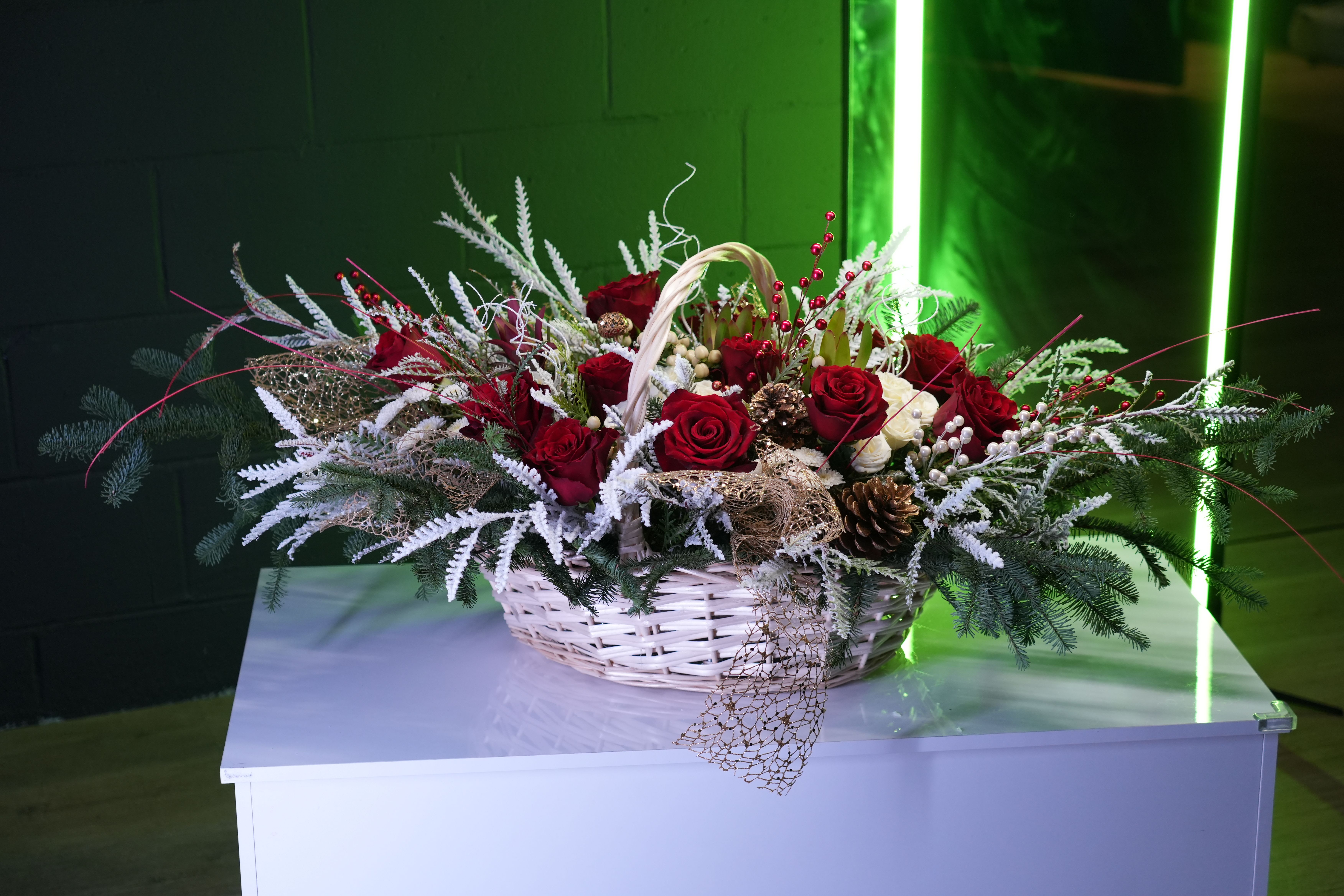 Festive Elegance Basket - The Festive Elegance Basket is a luxurious centerpiece designed to bring holiday cheer and sophistication to any celebration. Featuring velvety red roses, white accent blooms, and festive pinecones, this arrangement is enhanced with frosted branches, evergreen sprigs, and sparkling golden accents. Nestled in a charming white wicker basket, it is adorned with red berries and decorative elements, making it a stunning addition to holiday decor or a thoughtful seasonal gift. This arrangement embodies the warmth and beauty of the festive season.