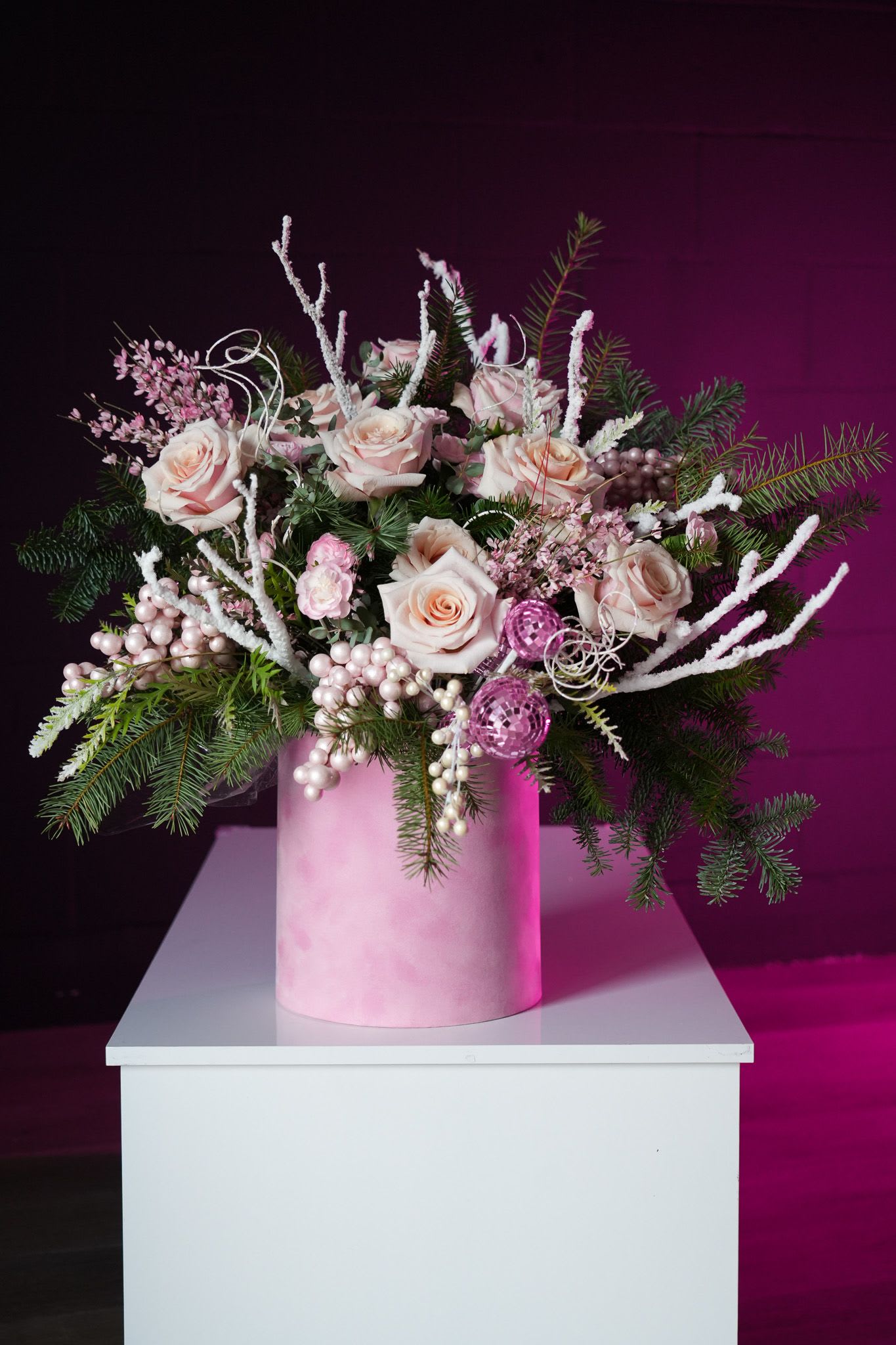 Winter Rose Splendor - The Winter Rose Splendor is a captivating arrangement that combines the delicate beauty of blush pink roses with the festive charm of winter greenery. Accentuated by frosted branches, shimmering pearl-like ornaments, and pink decorative accents, this arrangement is perfect for adding elegance to the holiday season. Arranged in a soft pink velvet hatbox, this piece exudes sophistication and seasonal magic, making it an ideal centerpiece or a luxurious gift for loved ones during the winter festivities.