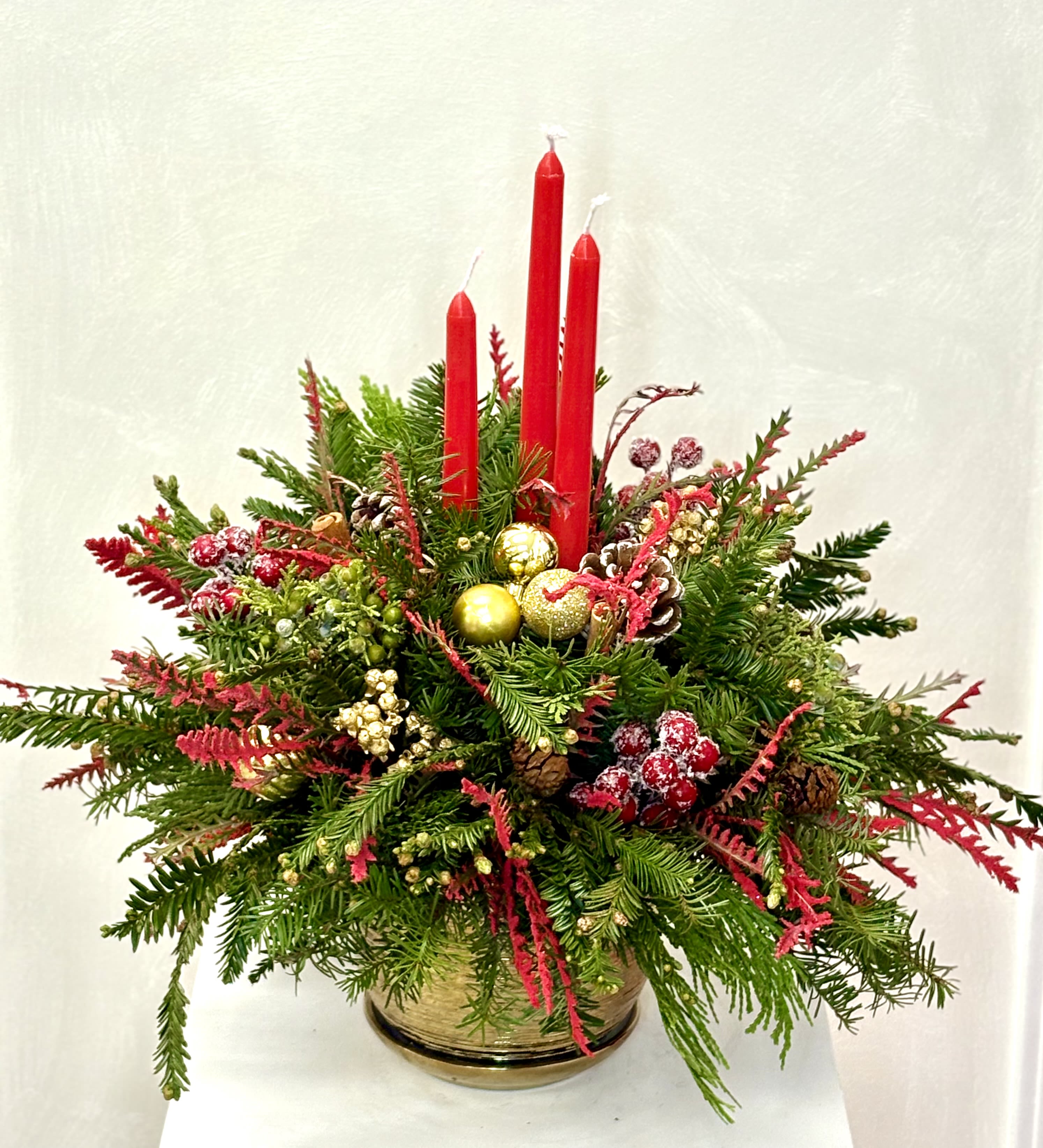Golden Noel Candle Glow By VLM Flowers - A festive holiday arrangement featuring lush evergreen branches accented with vibrant red candles, golden ornaments, pinecones, and winter berries. Designed in an elegant gold vase, this piece brings warmth, tradition, and seasonal sparkle—perfect as a Christmas centerpiece or a refined holiday gift.