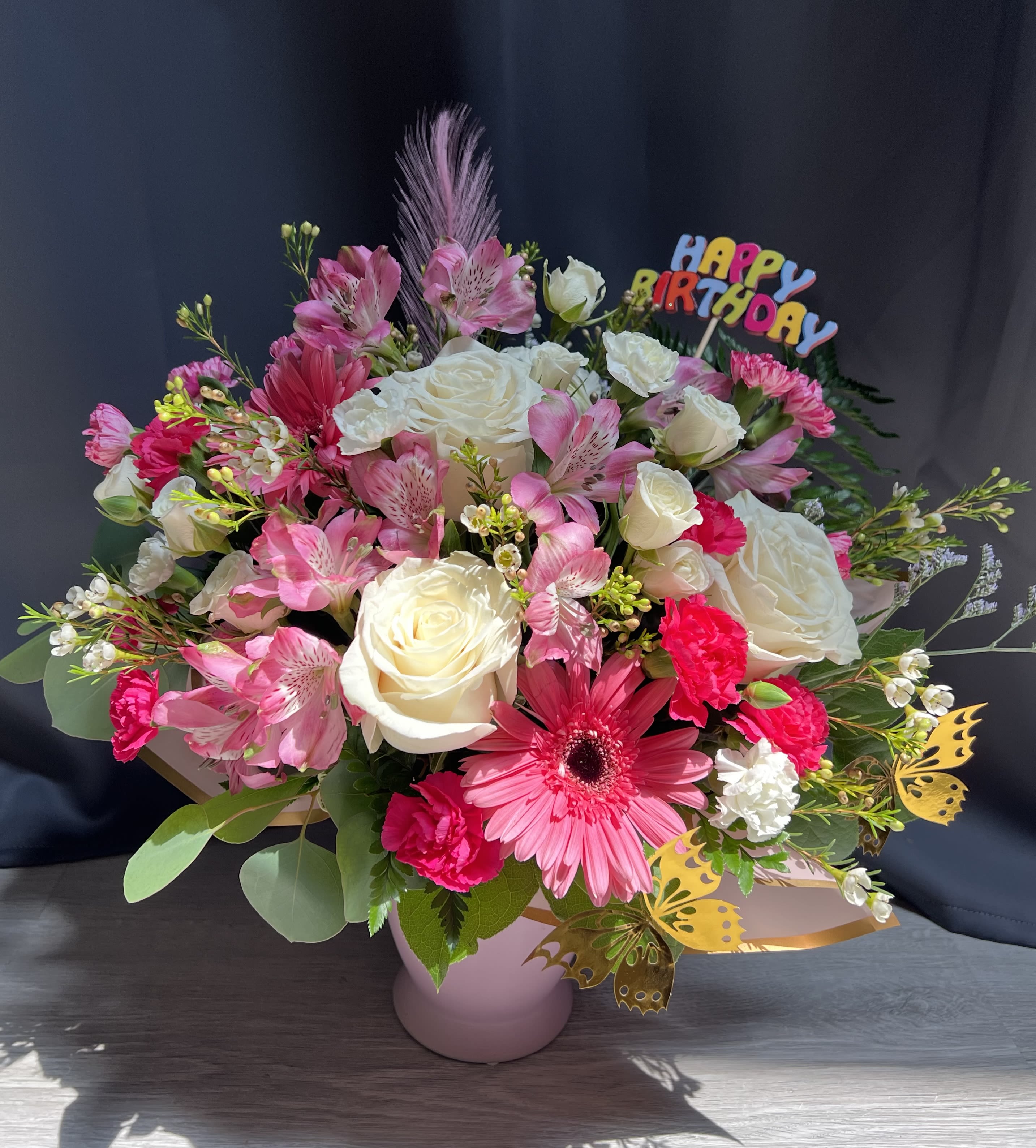 Pretty in Pink - Celebrate in full colour with this joyful floral arrangement, designed to brighten any birthday. Featuring a lush mix of creamy white roses, playful pink alstroemeria, vivid gerbera daisies, and cheerful carnations, this design bursts with vibrant textures and hues.   Accented with seasonal blooms, golden butterflies, and a festive “Happy Birthday” topper, it arrives beautifully presented in a stylish pink container. A perfect surprise for someone special, full of charm, colour, and celebration. 