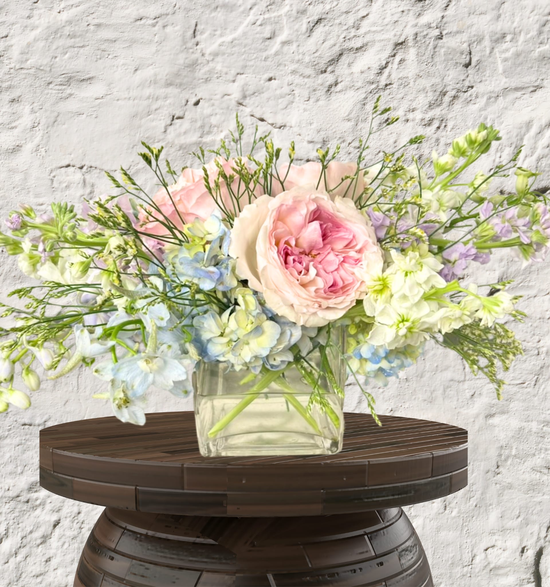Serene Sky Garden - Ethereal beauty inspired by a breezy afternoon.  This arrangement offers a refreshing, airy aesthetic that feels both modern and romantic. Centered around a lush, many-petaled pink Rose, the design features a soft palette of creamy white Stock and sky-blue Delphinium. Delicate Hydrangea and wispy greenery create a horizontal, "garden-style" silhouette that feels natural and unforced.  Housed in a glass vase this design is perfect for adding a sense of calm and sophistication to a coffee table, desk, or entryway.  Vibe: Fresh, airy, and calming.  Key Blooms:  Rose, Delphinium, and Stock.  Perfect For: Get well wishes, office decor, or a thoughtful housewarming gift.