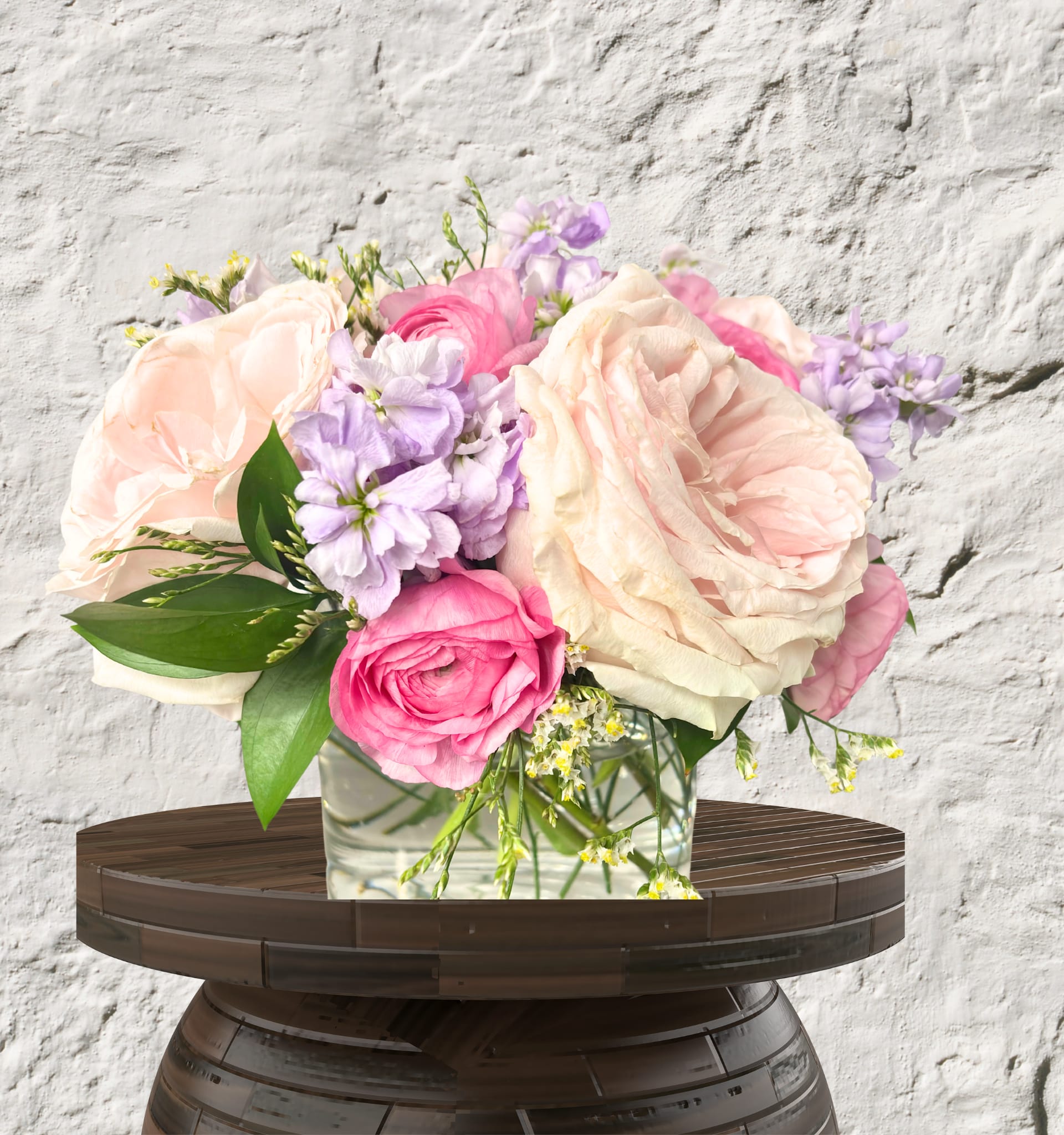 Blushing Blooms - Capture the romance of a secret garden with this lush, compact arrangement. This design features premium, many-petaled Roses and vibrant Ranunculus layered with fragrant Lilac Stock and delicate greenery.  Set in a sleek glass cylinder, its low-profile silhouette makes it an ideal centerpiece for intimate dinners or a sophisticated accent for a bedside table.  Vibe: Romantic, soft, and sophisticated.  Key Blooms:  Roses, Ranunculus, and Stock.  Perfect For: Anniversaries, "just because," or elevated home decor.