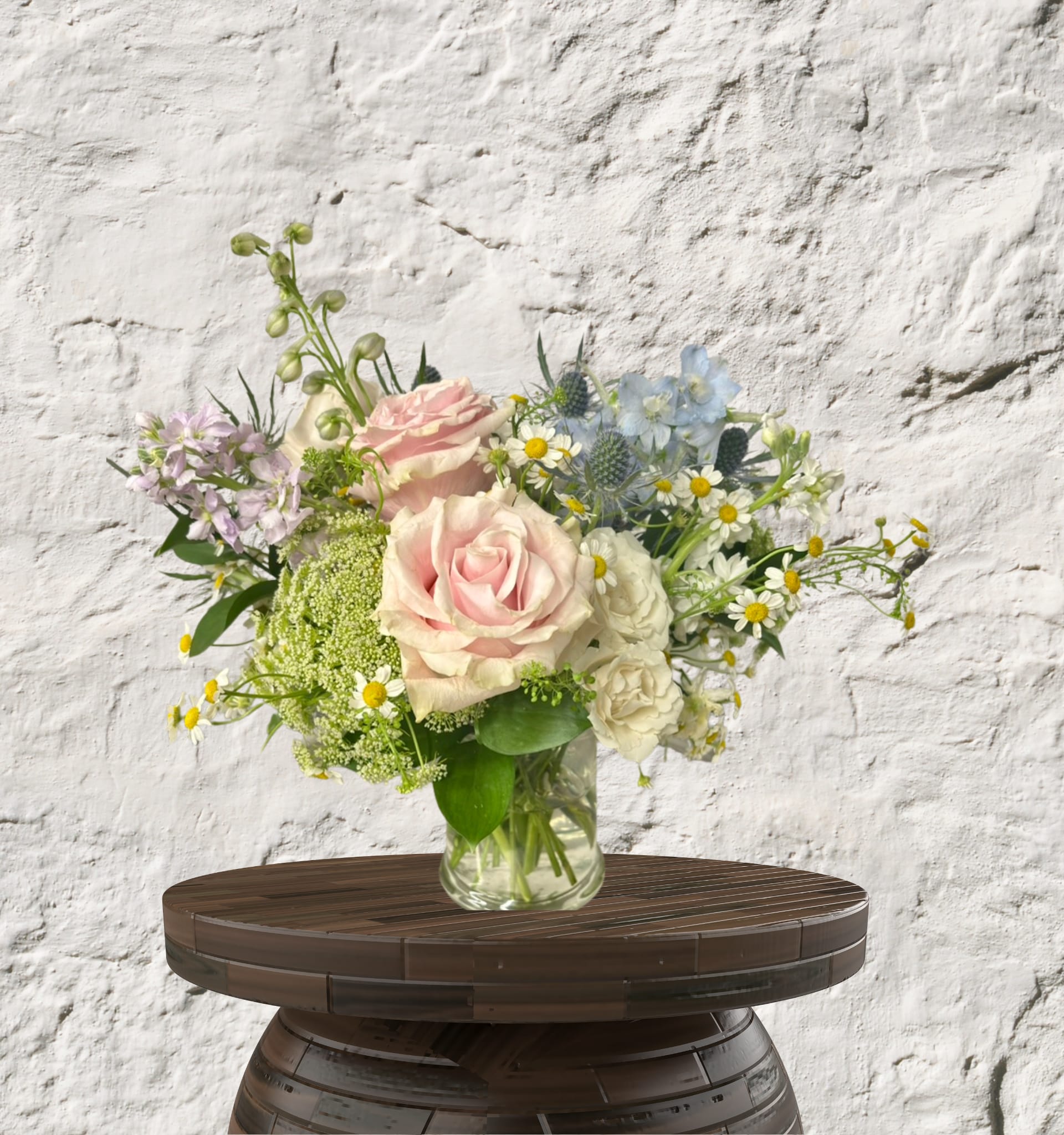 Blush Lyric - The Blush Lyric is a masterclass in "Petite Luxe" design, proving that a smaller footprint can still command a room. This arrangement is a sophisticated study in soft, airy color theory, featuring a dreamlike palette of blush pink, cream, and sky blue. It is designed for the modern connoisseur who appreciates intricate textures and a calm, artistic aesthetic.  The Design &amp; Florals This "Designer's Choice" edit features a multi-dimensional silhouette that encourages the eye to wander through layers of soft blooms and seasonal textures:  The Focal Heart: Large, blush pink roses anchor the arrangement, surrounded by ruffled cream carnations and delicate white stock for a romantic, garden-inspired core.  The Sky-Blue Edge: Striking spikes of vibrant blue delphinium provide architectural height and a daring contrast to the softer, muted tones.  Textural Whimsy: Artfully tucked throughout are sprigs of dusty miller, silver-toned foliage, and unique textural "spikes" that add a modern, high-end finish.  The Upscale Vessel: Presented in a heavy-weighted, clear glass cylinder, the minimalist foundation allows the complex play of stems and colors to be the star of the show.  