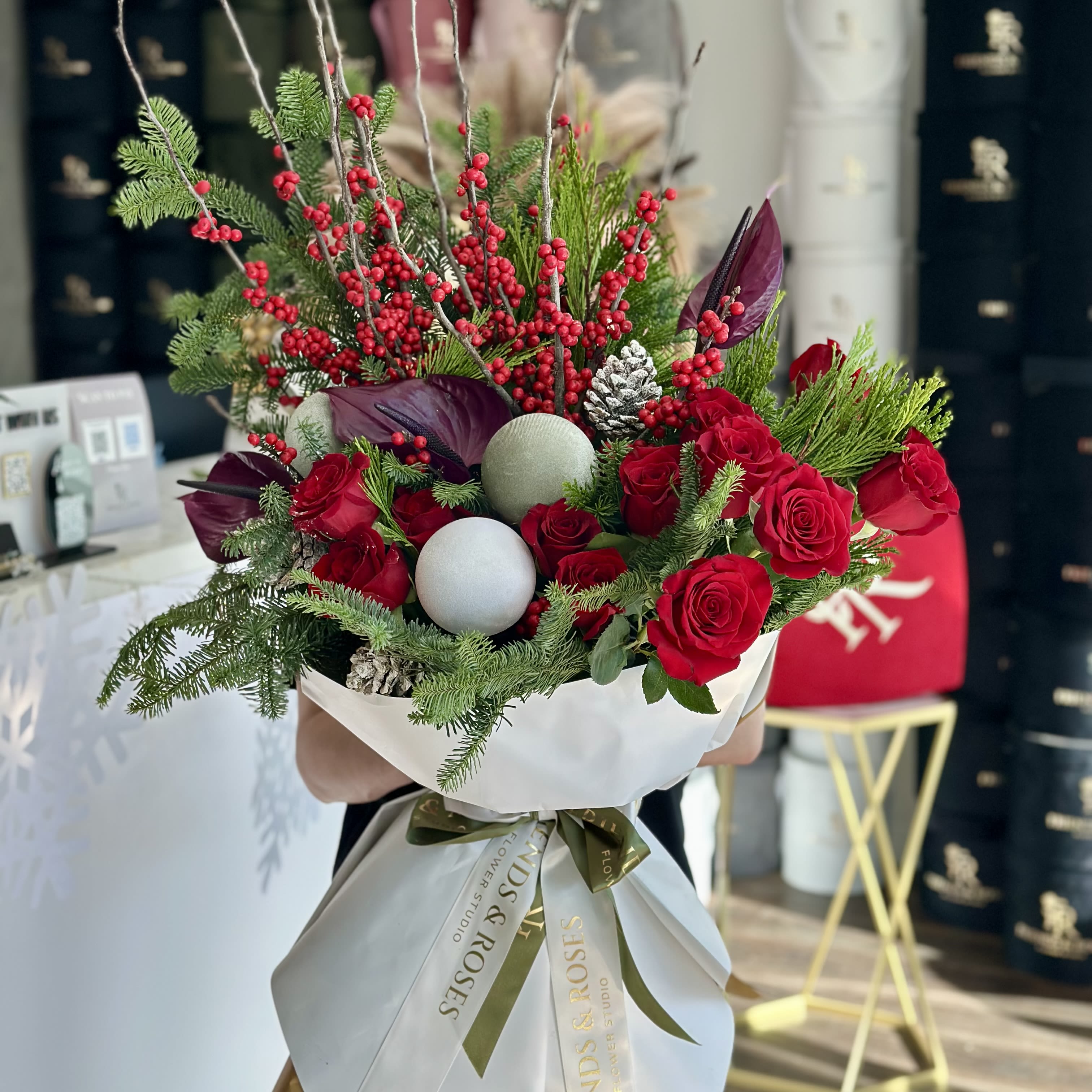 Winter Berry Elegance - A festive winter bouquet combining classic red roses with vibrant berry branches, rich anthurium blooms, and fresh evergreen foliage. Soft metallic ornaments and pine accents add a seasonal sparkle, creating a refined yet warm holiday statement.