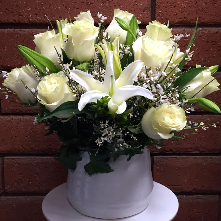 Winter in the City - Winter in the City – White Roses &amp; Lilies Arrangement  The charm and enchantment of New York City shine brightest in winter, and this elegant floral arrangement captures that magic beautifully. Winter in the City features premium Ecuadorian white roses paired with graceful Crystal Blanca lilies and airy genestra, all hand-arranged in a sleek white ceramic container. The crisp whites and soft textures evoke snowy streets, glowing city lights, and classic winter sophistication.  Perfect for winter celebrations, sympathy gestures, or refined seasonal décor, this arrangement brings calm beauty and modern elegance to any space.  • Standard - 9 Luxury White Roses, 3 Stems of Crystal Blanca Lilies, Genestra and Fresh Garden Greens in White Ceramic Vase (Approximate size: 10" W x 11" H)  • Deluxe - 12 Luxury White Roses, 4 Stems of Crystal Blanca Lilies, Genestra and Fresh Garden Greens in White Ceramic Vase  • Premium - 15 Luxury White Roses, 5 Stems of Crystal Blanca Lilies, Genestra and Fresh Garden Greens in White Ceramic Vase  Please Note: Lilies are typically delivered in bud form to ensure maximum freshness and longevity. As blooms open, gently remove the stamens to prevent pollen staining and extend the life of the flowers.  Bring home the serene elegance of winter with this New York–inspired floral arrangement, designed to enchant all season long.