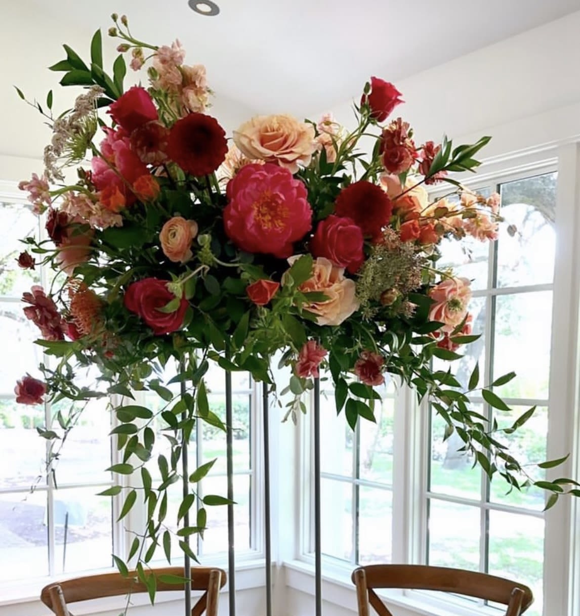 Grand Estate Elevated Centerpiece - The Grand Estate Elevated Centerpiece Make a breathtaking statement with our Grand Estate Elevated Centerpiece, designed to bring high-impact drama and vertical scale to your reception. By lifting the floral artistry above the table, this arrangement creates a "ceiling of flowers" that fills the room with luxury while keeping sightlines clear for guest conversation.  Design &amp; Artistry Architectural Height: Mounted on elegant, slender risers or glass pedestals, these arrangements draw the eye upward to appreciate the full volume of your venue.  Lush &amp; Overflowing: Features a generous mix of premium focal blooms, such as hydrangea and long-stemmed roses, accented with cascading greenery for a romantic, weeping effect.  360-Degree Design: Expertly crafted to look spectacular from every angle, ensuring a consistent, high-end experience for every guest at the table.  Custom Stylings: Fully tailored to your wedding’s aesthetic, whether you prefer a structured, classic dome or a wild, airy garden look.  The Grand Impact Tall centerpieces are the ultimate way to transform large ballrooms or high-ceilinged tents into an intimate, botanical sanctuary. This design provides an editorial, "wow-factor" moment that defines your reception space with unparalleled elegance and sophistication.