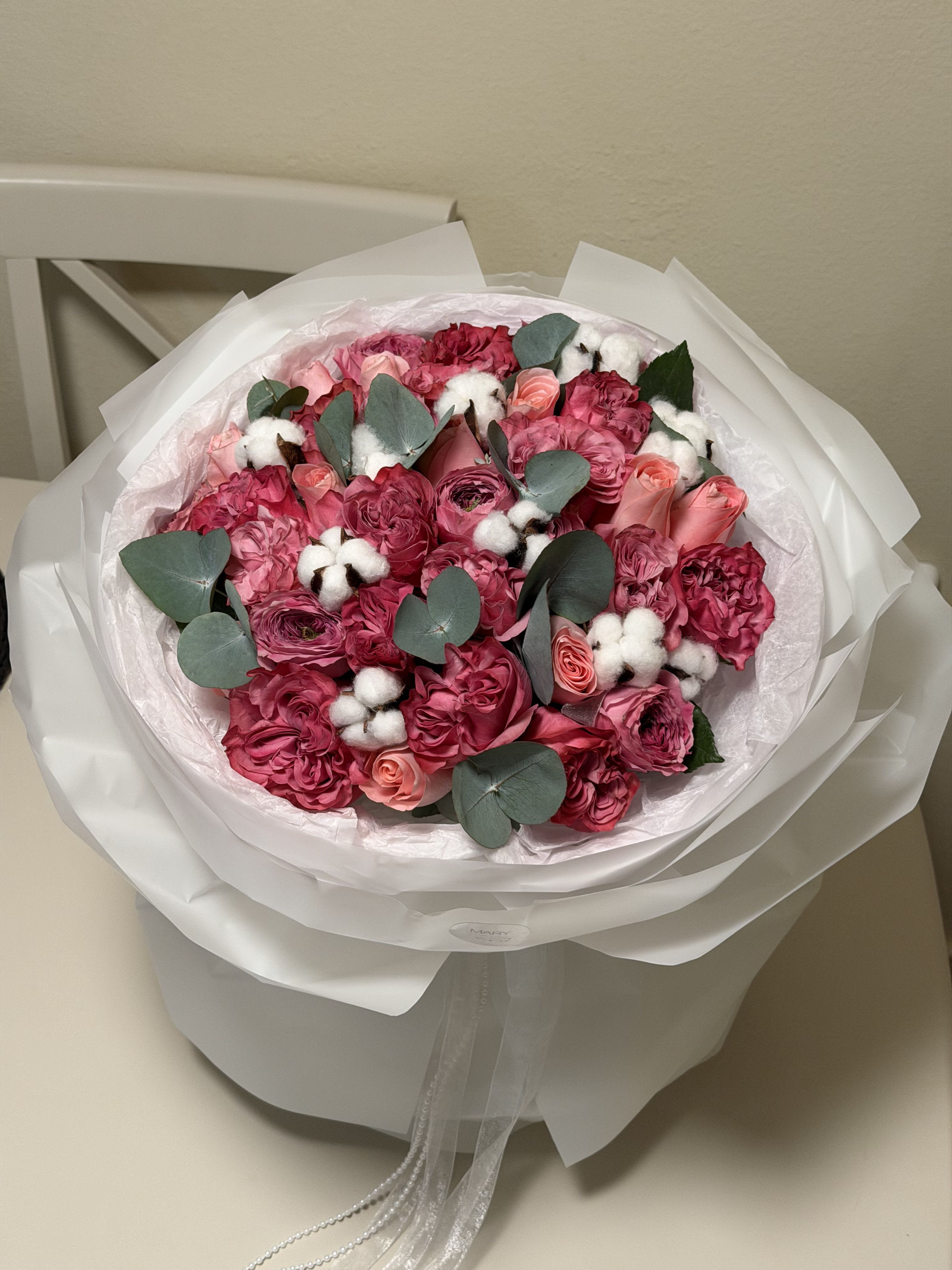 Pink Symphony Arrangement  - This exquisite bouquet is a true statement of elegance and romance. Featuring lush shades of deep rose, blush pink, and soft peach blooms, it is beautifully complemented with delicate cotton accents and fresh eucalyptus greenery. Expertly arranged and wrapped in premium white packaging, this bouquet brings a luxurious and refined touch to any occasion. 